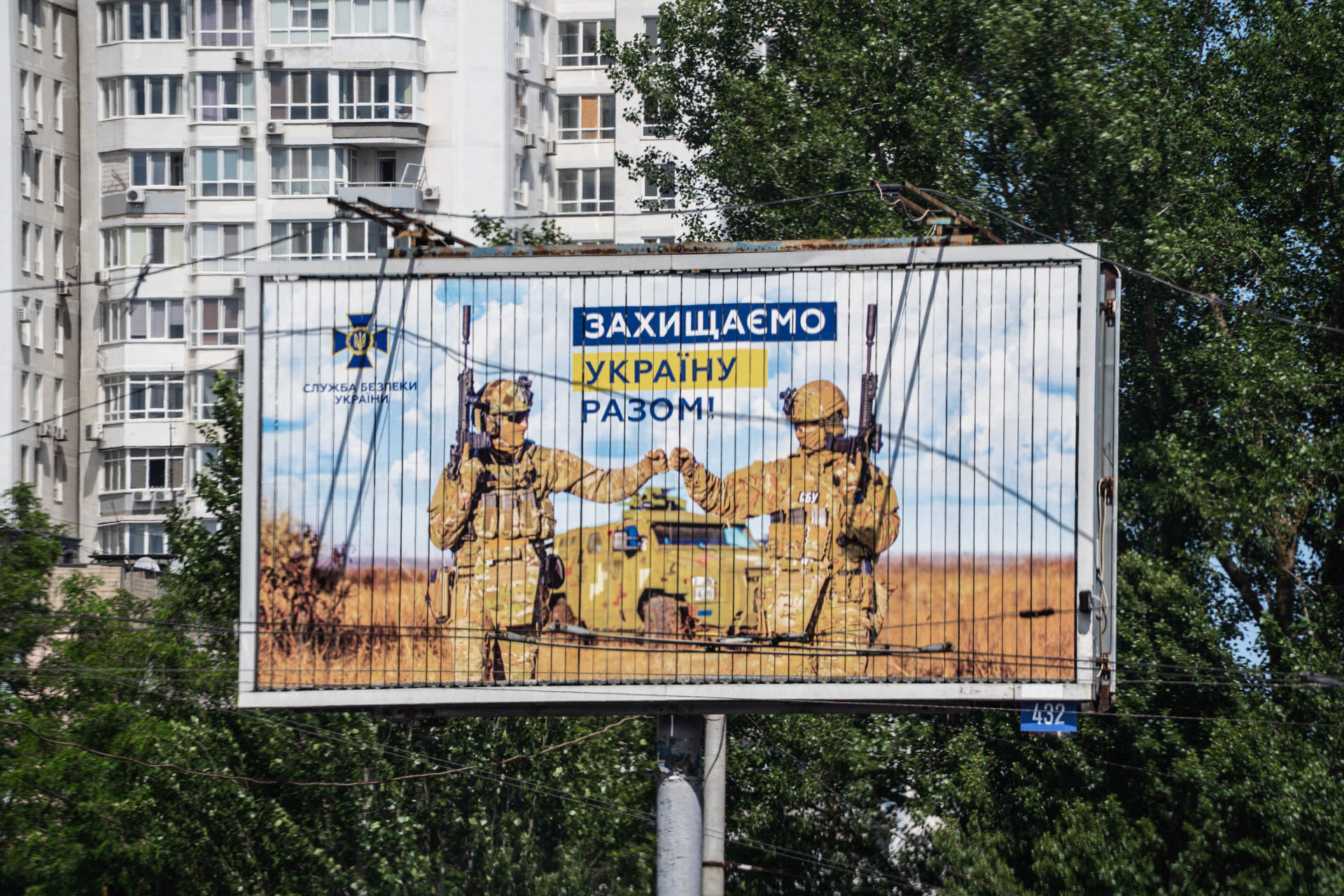 A billboard by the Security Service of Ukraine (SBU) reading "Захищаємо Україну разом!" (Defending Ukraine Together!) stands in front of a residential building in Odesa, Ukraine, on [June 12, 2026]. The poster shows two servicemen bumping fists in front of an armored vehicle in a wheat field.