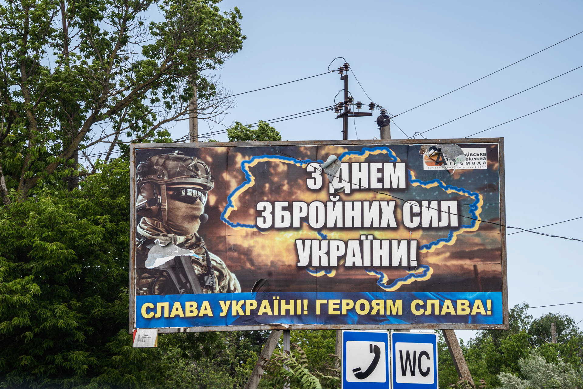 A billboard marking the Day of the Armed Forces of Ukraine, reading "З Днем Збройних Сил України!" (Happy Day of the Armed Forces of Ukraine!) and "Слава Україні! Героям Слава!" (Glory to Ukraine! Glory to the Heroes!), stands near power lines and public signage in Odesa, Ukraine, on June 12, 2026. The poster depicts a soldier beside an outline of Ukraine against a battlefield background.