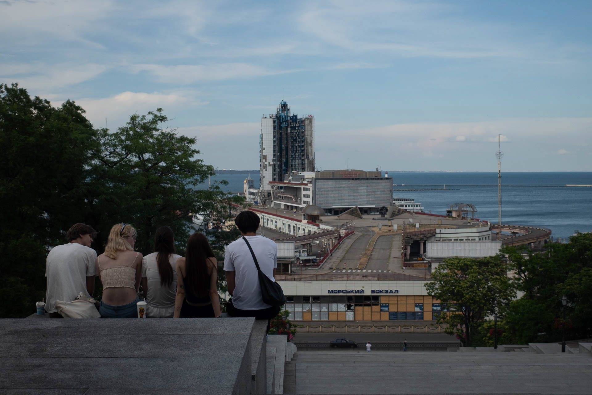 Five young people sit at the top of the Potemkin Stairs overlooking the port of Odesa, Ukraine, at dusk on June 12, 2026, with the war-damaged Hotel Odessa high-rise and the Maritime Terminal building in the background. The hotel, located at the sea terminal, was damaged during Russian attacks on the port of Odesa.
