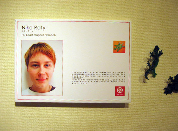 Exhibition wall panel with a portrait of designer Niko Räty and Japanese text about the PC Beast magnet/brooch, with three dark green plastic lizard-shaped pieces mounted on the wall to the right.