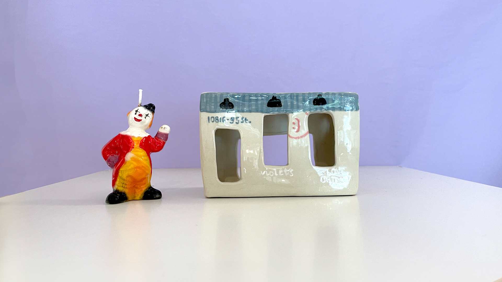 A small painted clown shaped candle and a small ceramic version of Violets' storefront,.