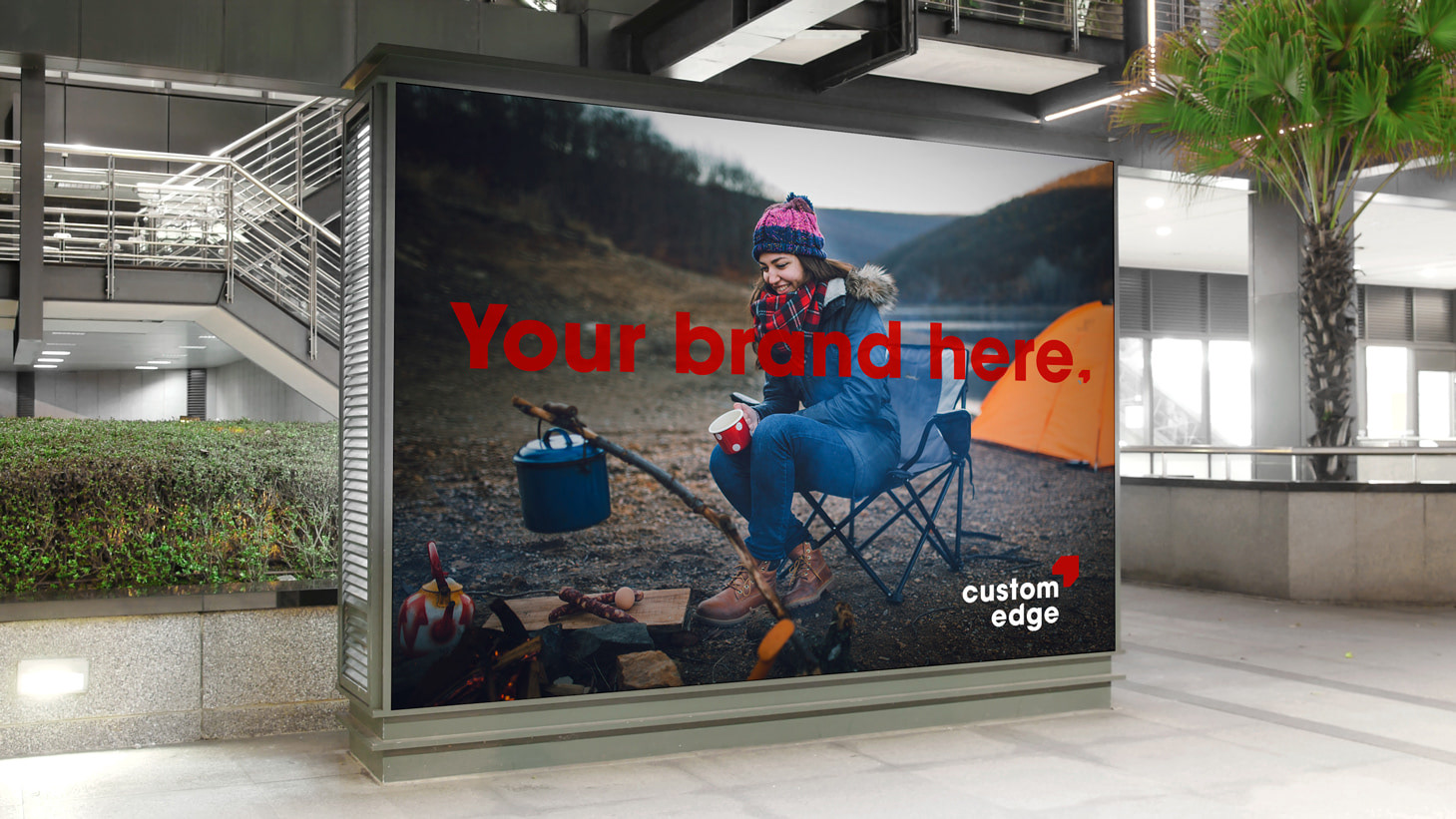 A Custom Edge indoor billboard ad with a photo of a woman sitting at a campsite. Text reading "Your brand here" is laid over top.