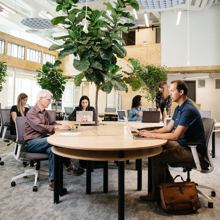 A bright co-working space with a diverse group of people sitting at a shared table working on laptops.