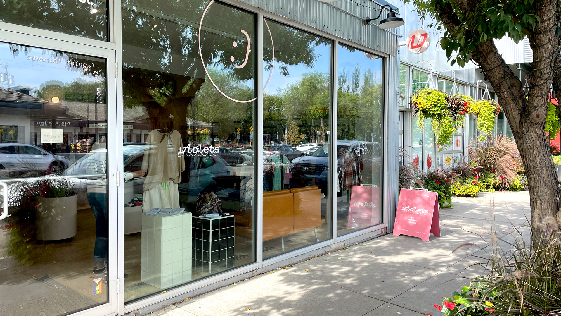 A sunny storefront of windows with the violets logo and large smiley face decal. Plants and trees line the sidewalk, alongside a pink violets sandwich board sign.