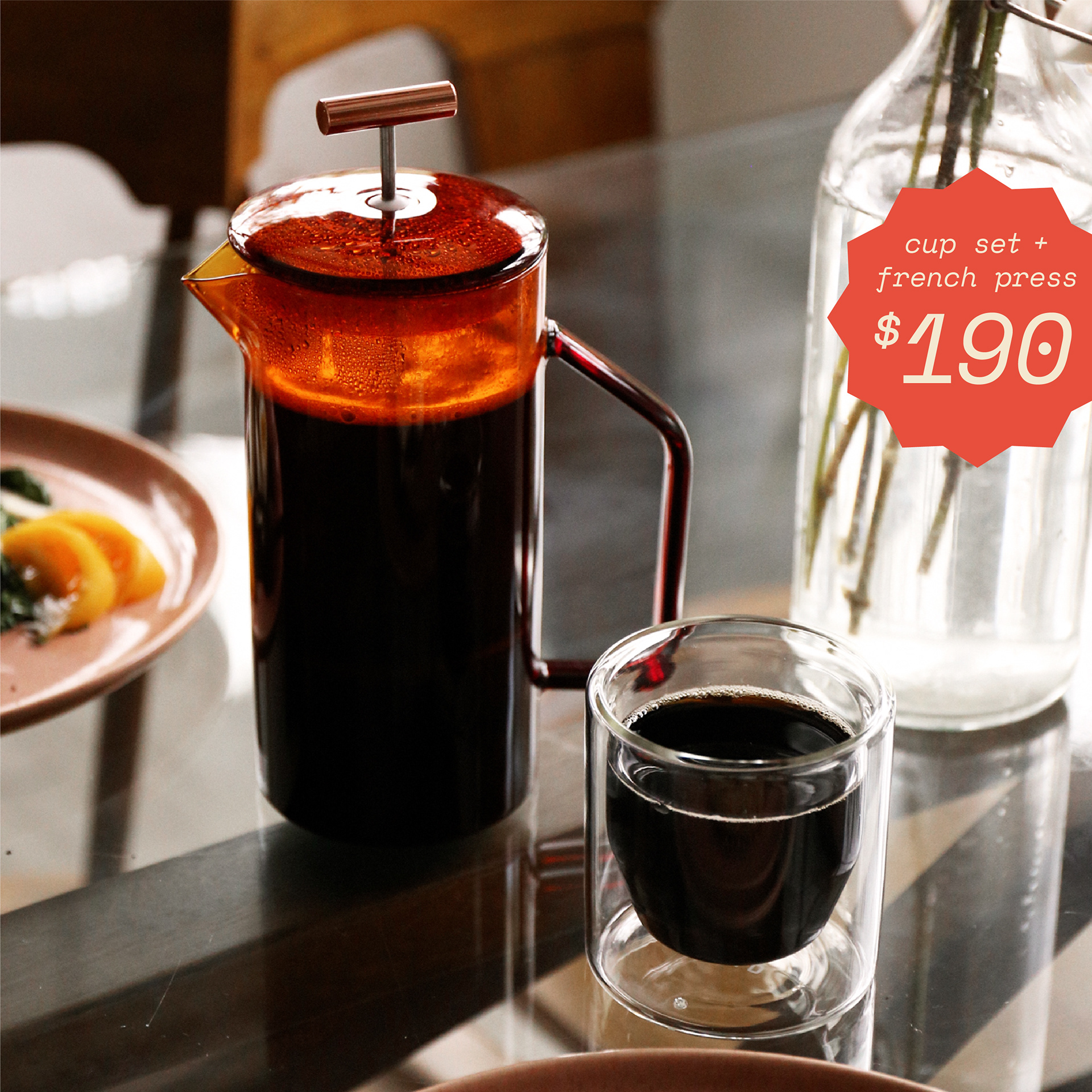 A table with an amber glass french press and a clear glass, double walled cup, both filled with coffeeA starburst shape containing text "cup set and french press 190 dollars"