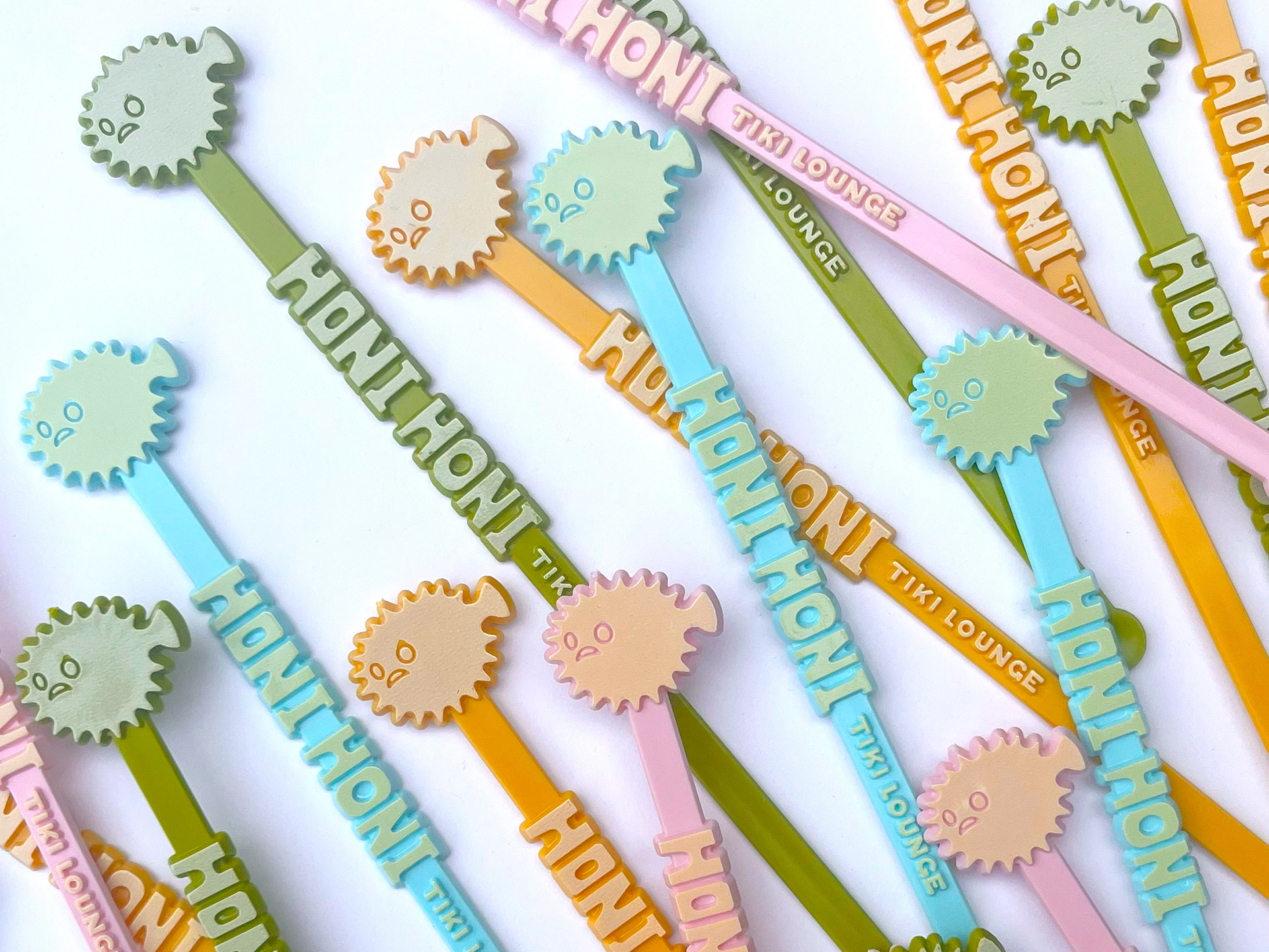 Custom-made Honi Honi swizzle sticks scattered on a white background. The sticks have a blowfish on the end, Honi Honi Tiki Lounge runs down the stick in 3D lettering.