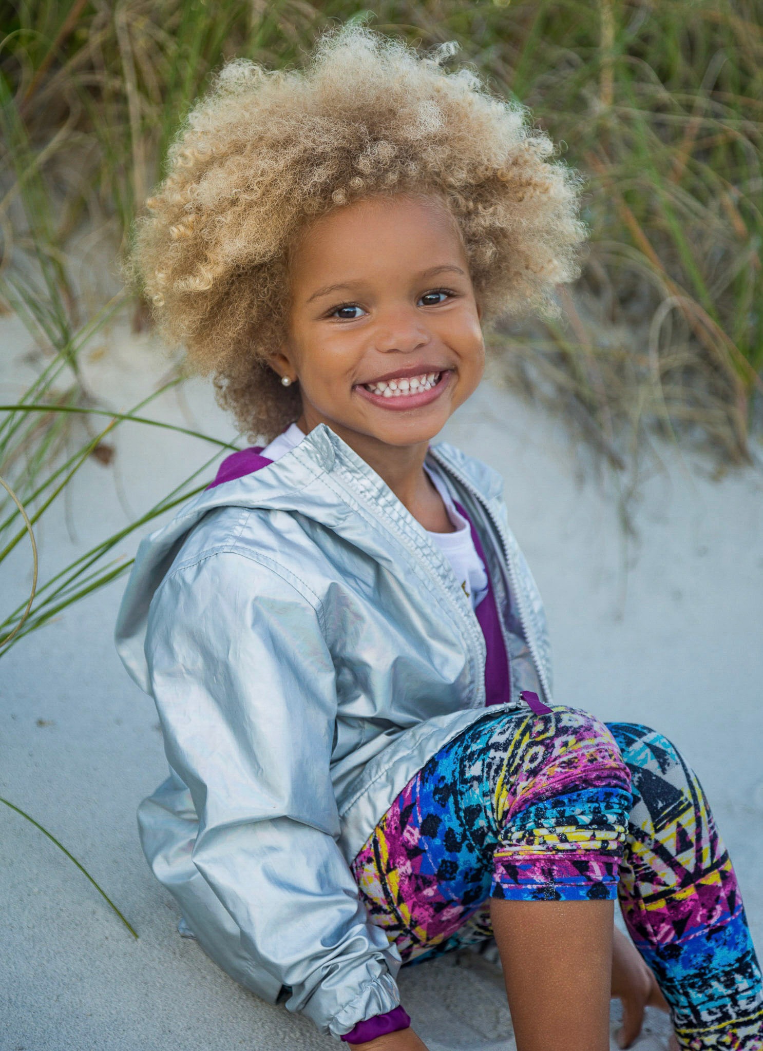 Smiling young girl with curly hair sitting on the sand dunes during Jacksonville Beach Family Photo Session