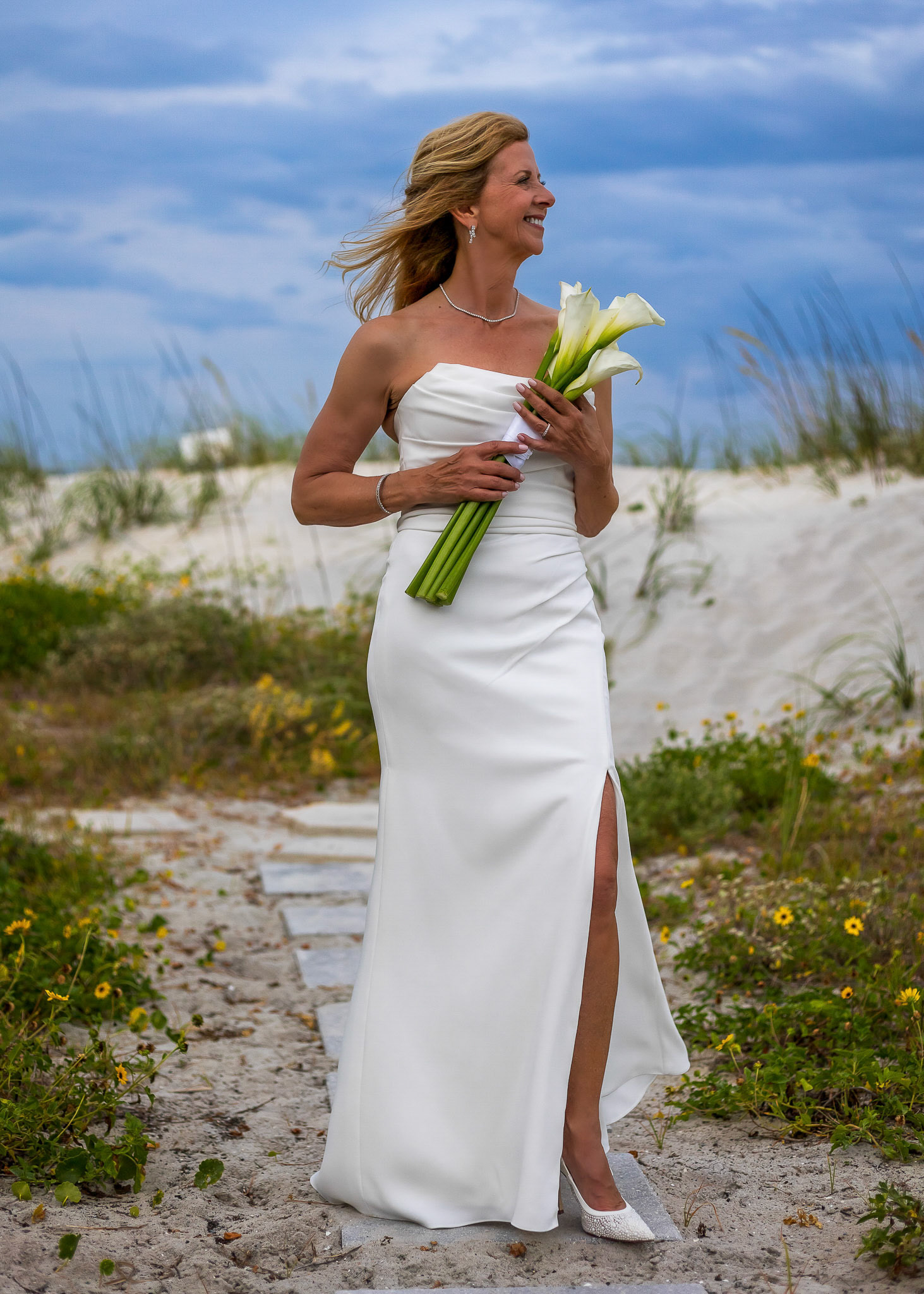 Radiant bride smiling on a breezy beach, wearing a white strapless gown and holding an elegant white calla lilies bouquet