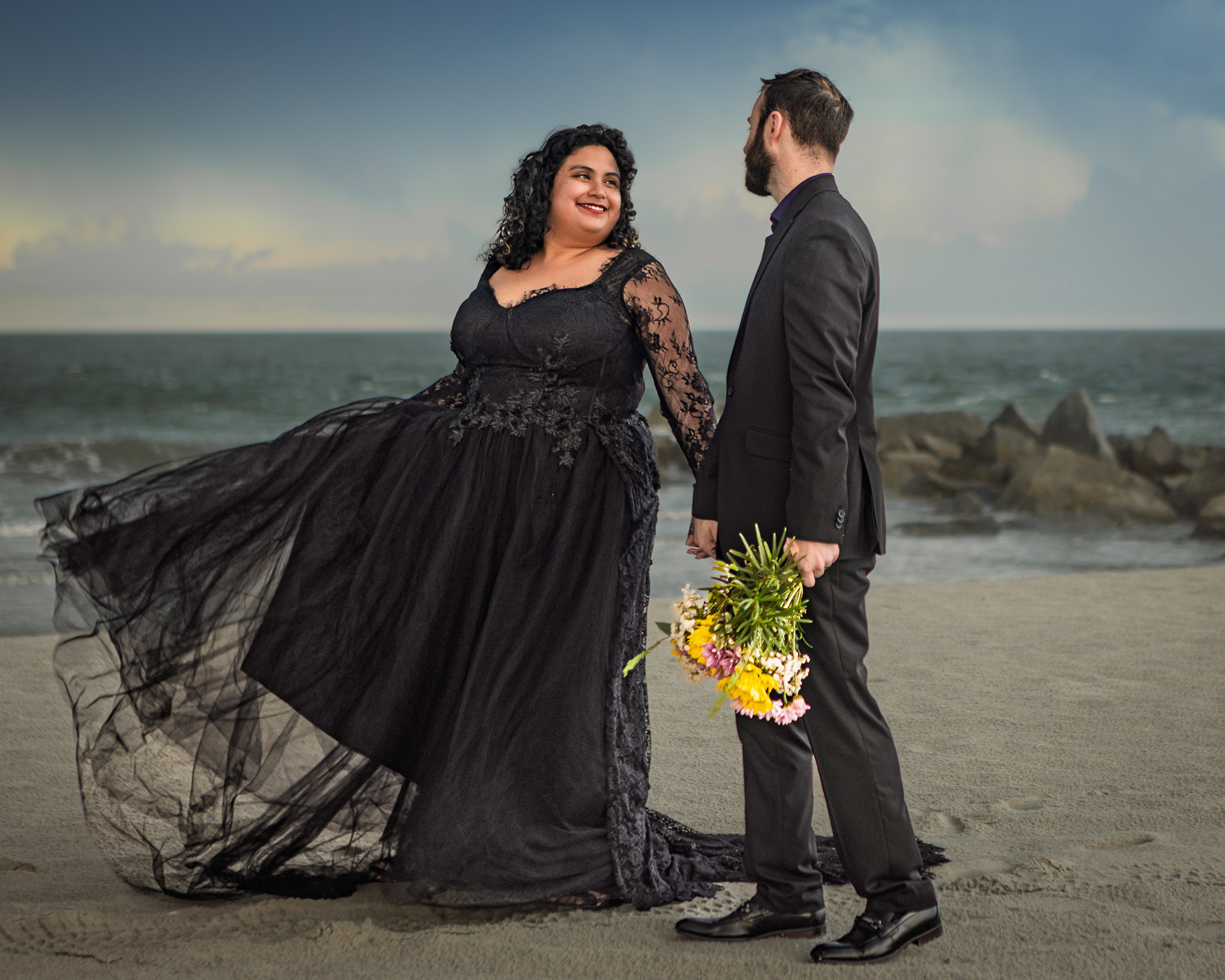 Cinematic wide shot of a couple in black wedding lace gown and suit walking on Villano Beach during stormy skies