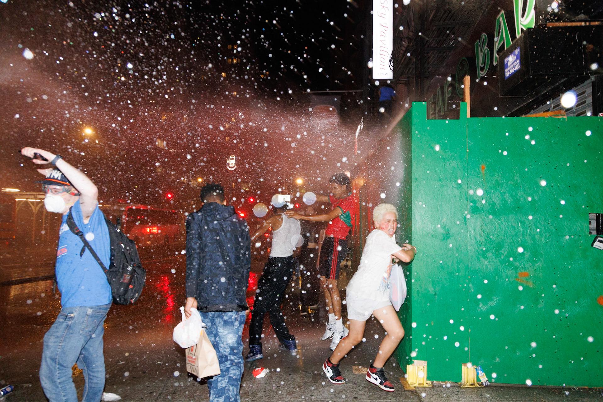 People Dodging Water Spray Near Green Bar Wall at Night