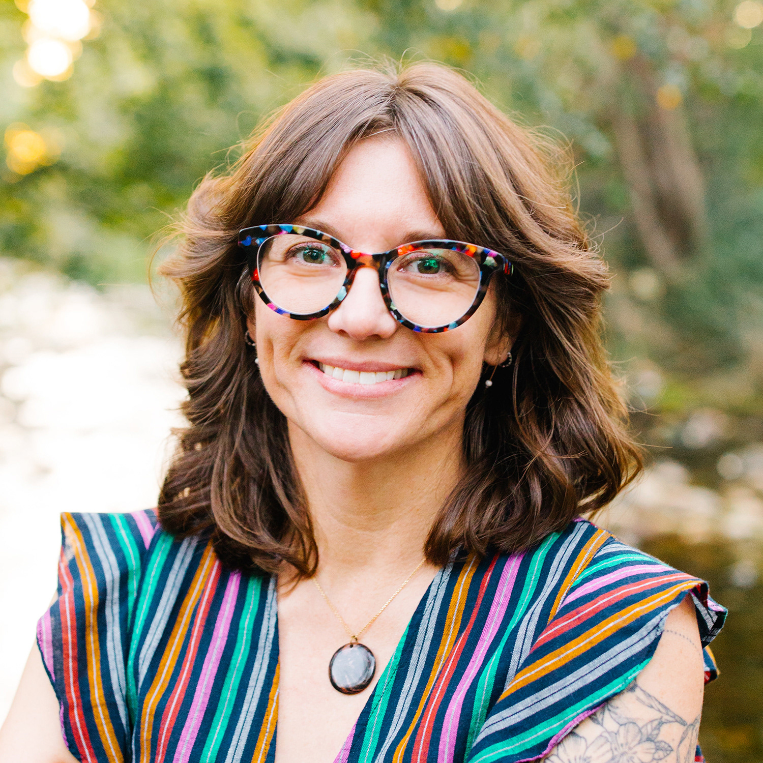 Headshot of Heidi M. Rogers, an author and illustrator of picture books, young adult, middle grade, and graphic novels. With a blurred background of green trees and a babbling brook, Heidi is a white woman with wavy brown hair and black glasses with rainbow tortoise-shell specks. Her arms are crossed over the front of her rainbow-striped dress and she has a warm and welcoming smile on her face.