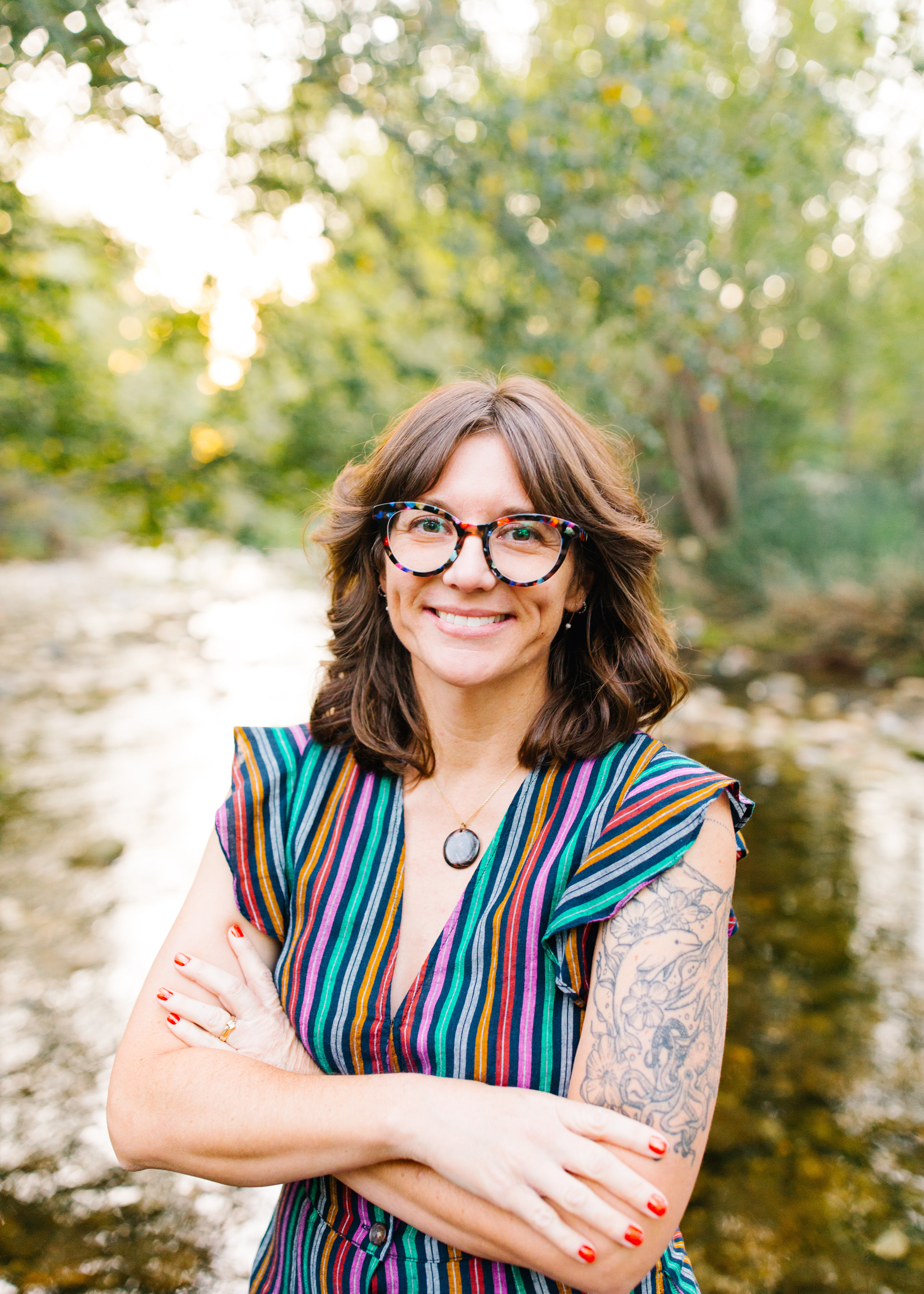 Headshot of Heidi M. Rogers, an author and illustrator of picture books, young adult, middle grade, and graphic novels. With a blurred background of green trees and a babbling brook, Heidi is a white woman with wavy brown hair and black glasses with rainbow tortoise-shell specks. Her arms are crossed over the front of her rainbow-striped dress and she has a warm and welcoming smile on her face.