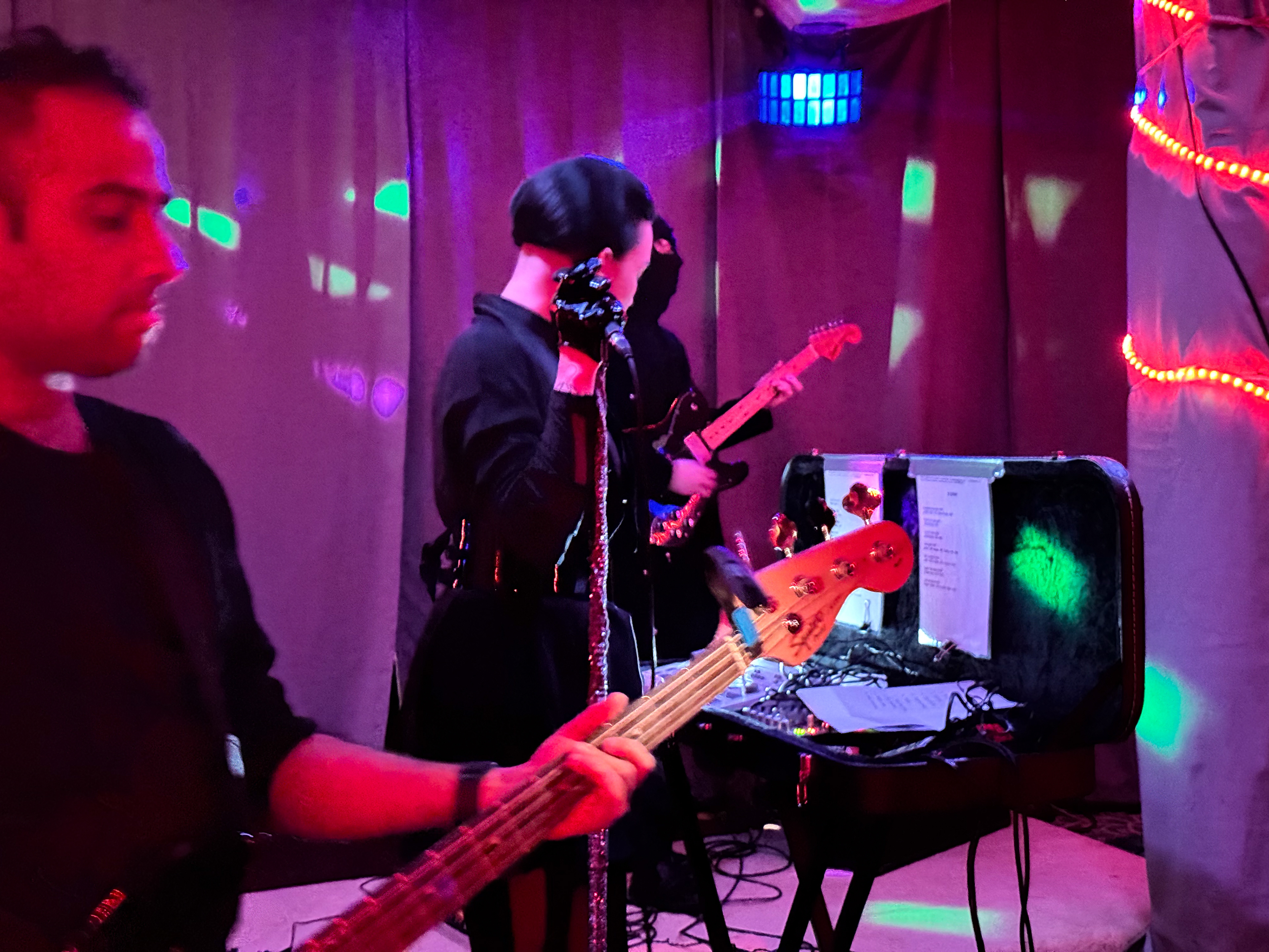 Faux Fear perform at Pleasuretone in Reading, PA.  All three members wear black clothing.  The male bass player appears from the left of the frame playing his instrument.  At the center of frame, the female vocalist looks into an open guitar case containing drum machines and a synthesizer.  Her right hand grips a microphone atop a stand wrapped in reflective material.  The light of the room reflects heavily from her glove.  The guitarist peers from behind her wearing a black mask obscuring his face.  The venue has curtains covering the walls and a column wrapped in orange LED string lighting is visible on the right of frame.  The lighting has very strong red and purple overtones, with patches of purple, blue, and green light projected across the room.