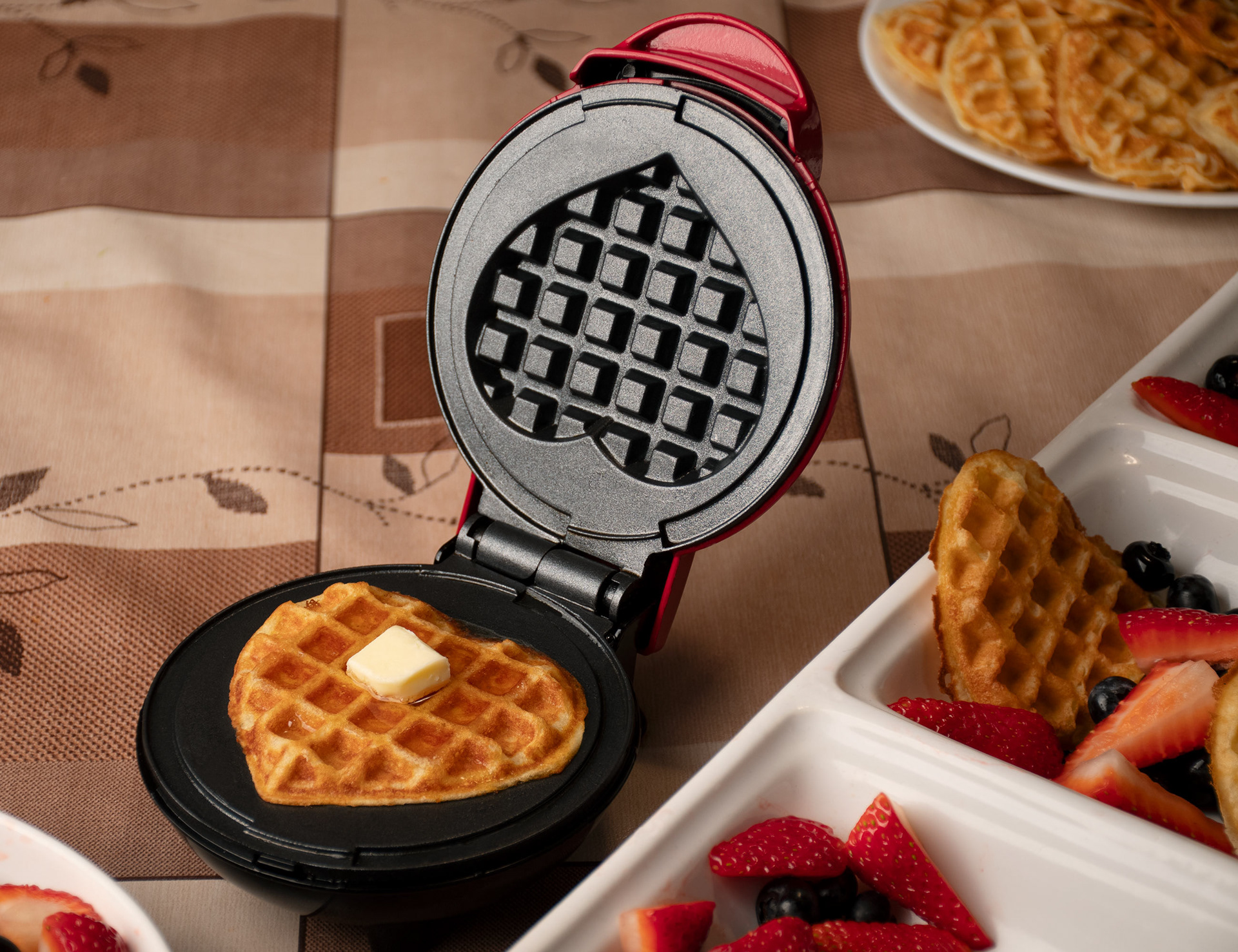 Waffle with butter and syrup on a red waffle maker with strawberries and blueberries on white plates on the sides