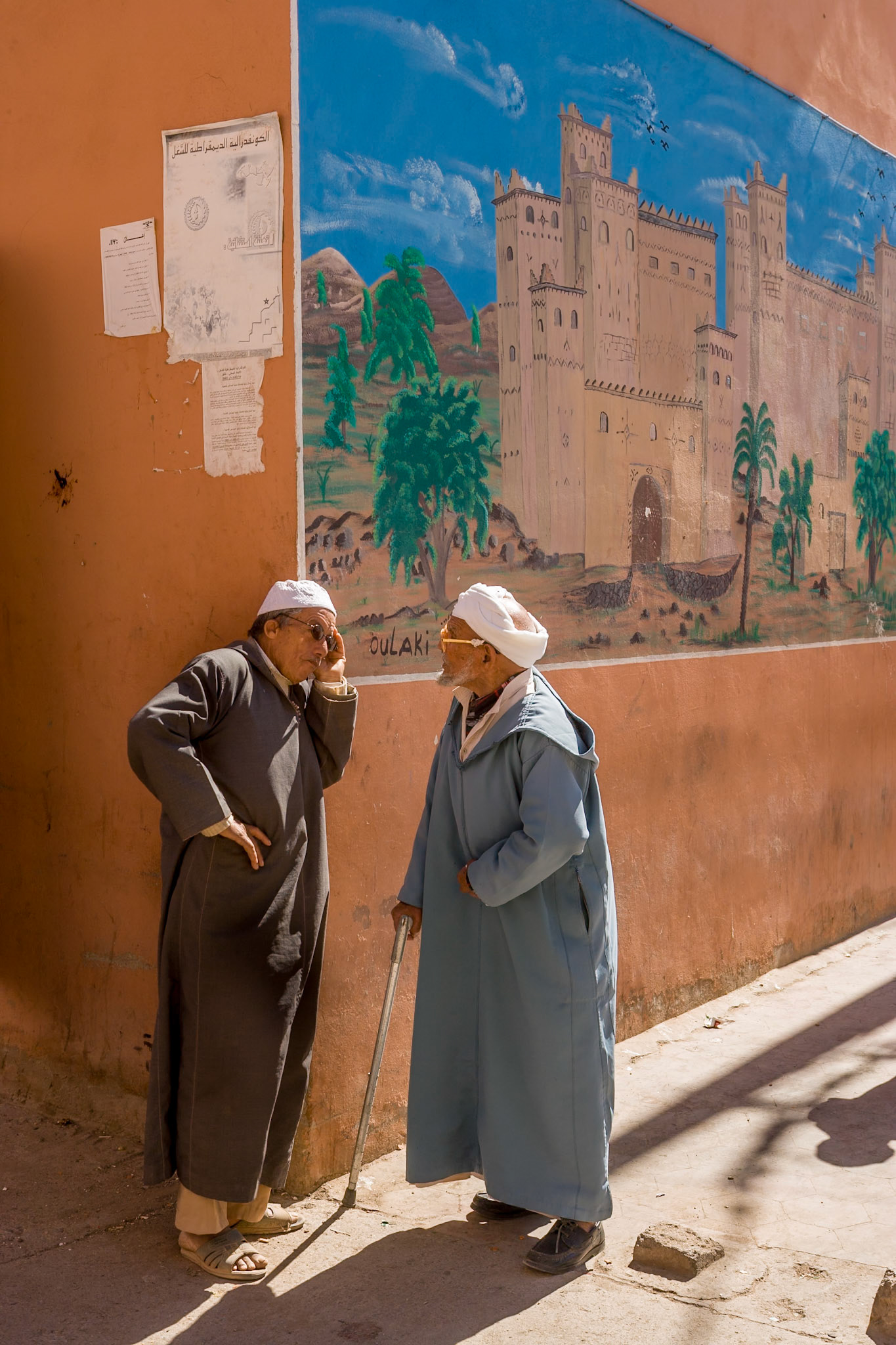 Two Man talking at the corner of the street where a big painting of a city wall is painted at Tinerhir