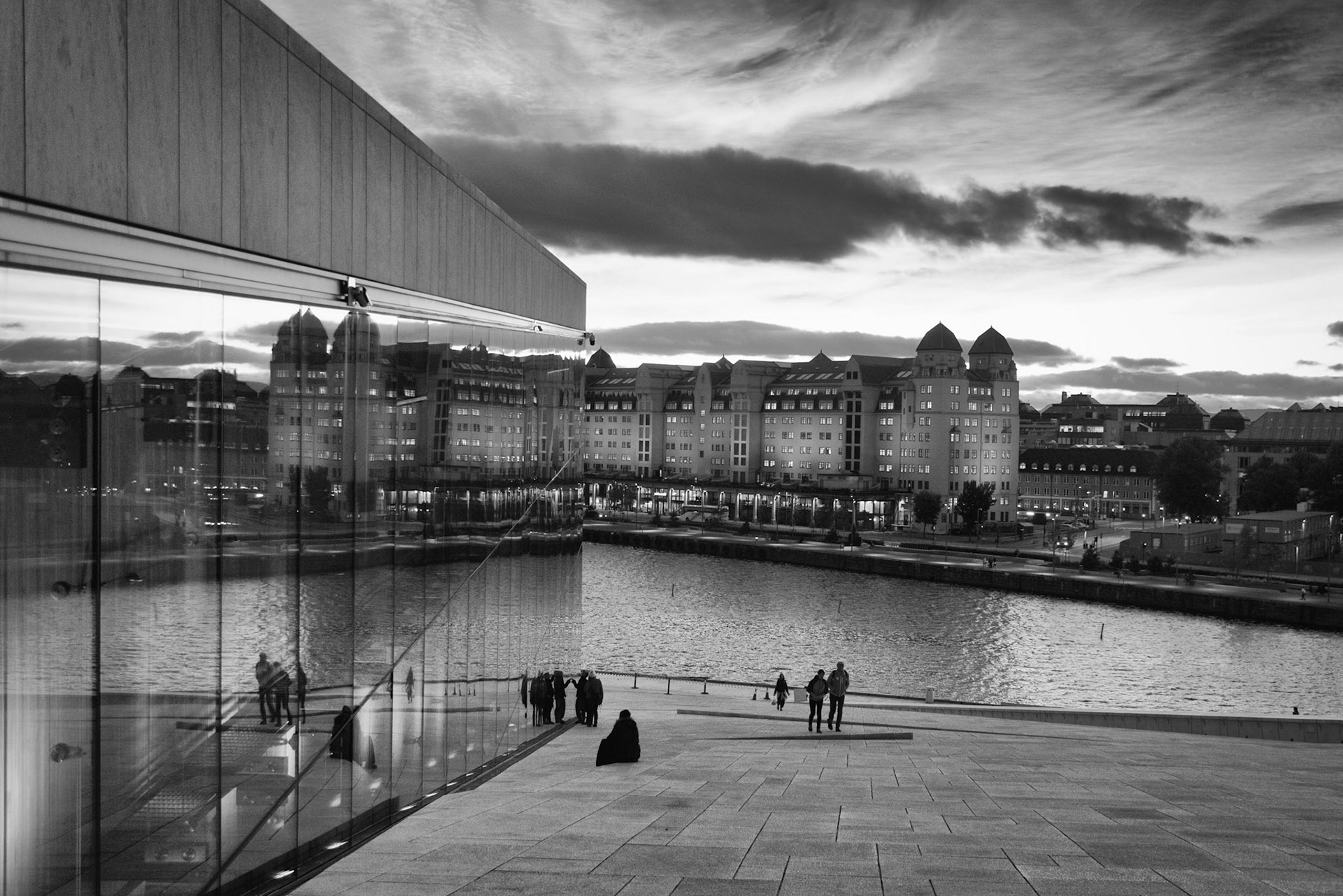 People walking on the Oslo Opera House and Havnelageret's reflection on the glass facade in Oslo, Norway