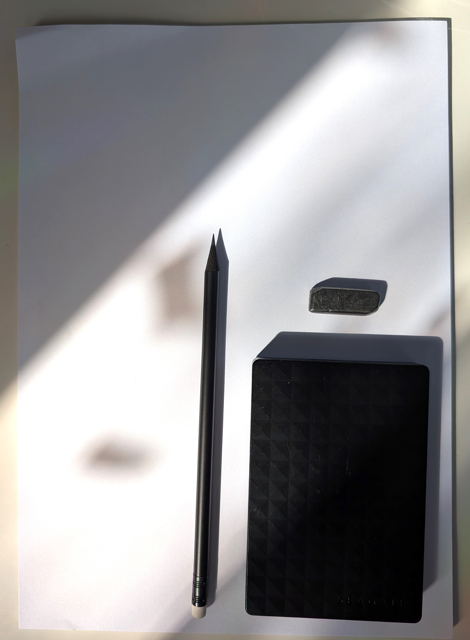 Ein weißes DIN A4 Blatt mit Sonnenlicht. Auf dem Papier liegen ein schwarzer Stift, eine schwarze externe Harddrive und ein Stück Graphit.