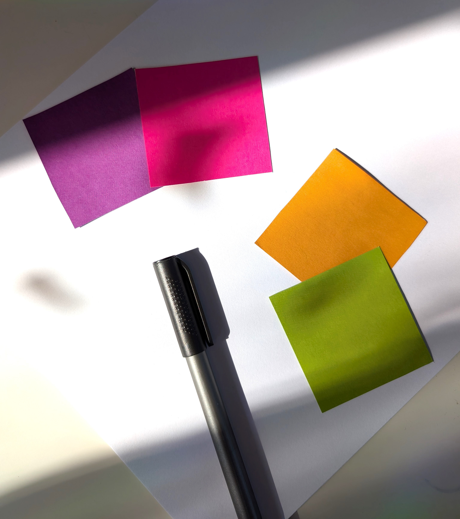 Ein weißes Papier, darauf liegen vier kleine quadratische Farbflächen.  Lila, Magenta, Orange und Grün. In der Mitte liegt ein schwarzer Stift.