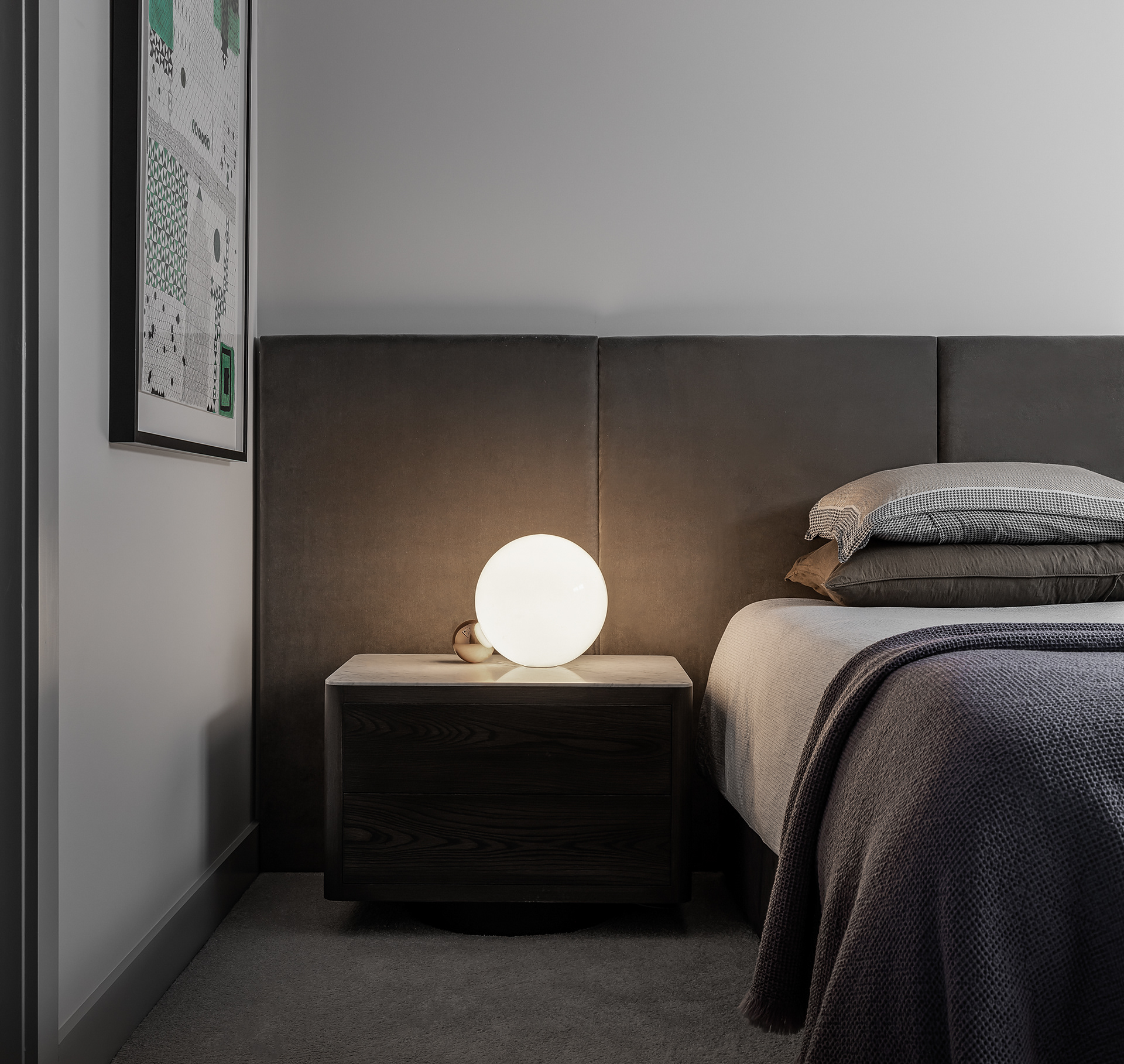 Bedroom interior with a grey upholstered headboard, layered neutral bedding and a glowing spherical bedside lamp.