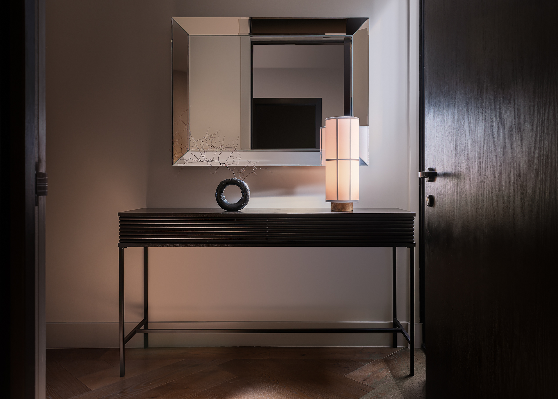 Dark console table with a sculptural circular vase and illuminated table lamp beneath a contemporary framed mirror.