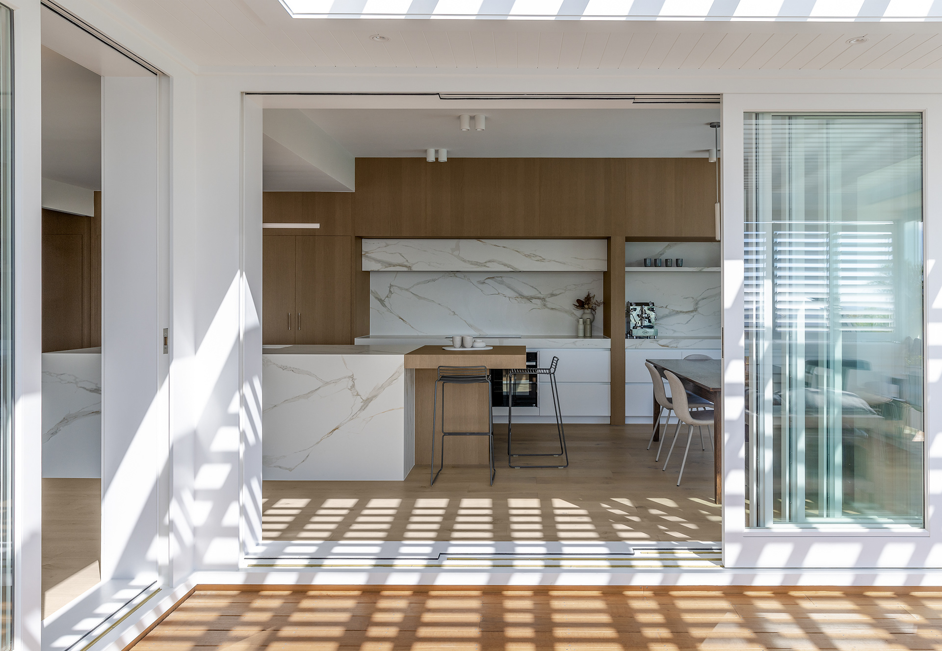 Codrington Residence kitchen viewed from the outdoor pergola, showing patterned sunlight across the open-plan interior