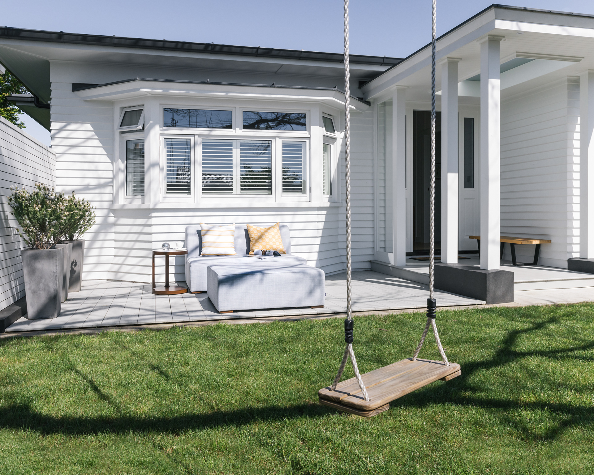 White weatherboard bungalow with outdoor lounge area and timber swing in a landscaped garden