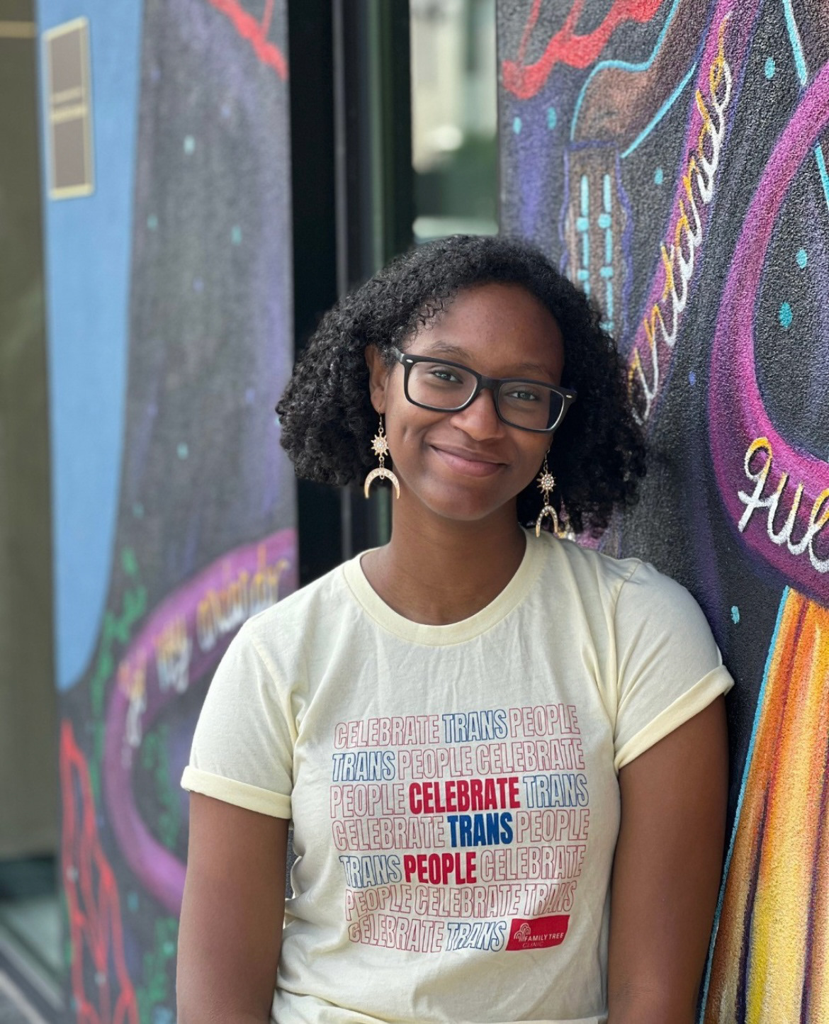 A person leans against a mural. She smiles and looks into the camera wearing a light yellow t-shirt that reads, celebrate trans people.