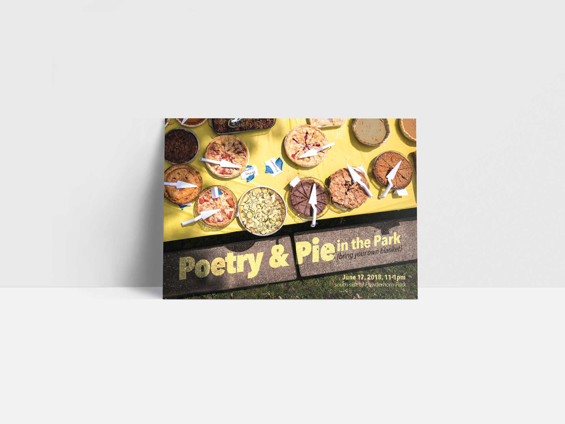 A postcard with a photograph of a park picnic table covered with different types of pies. The park bench reads, poetry and pie in the park, bring your own blanket.