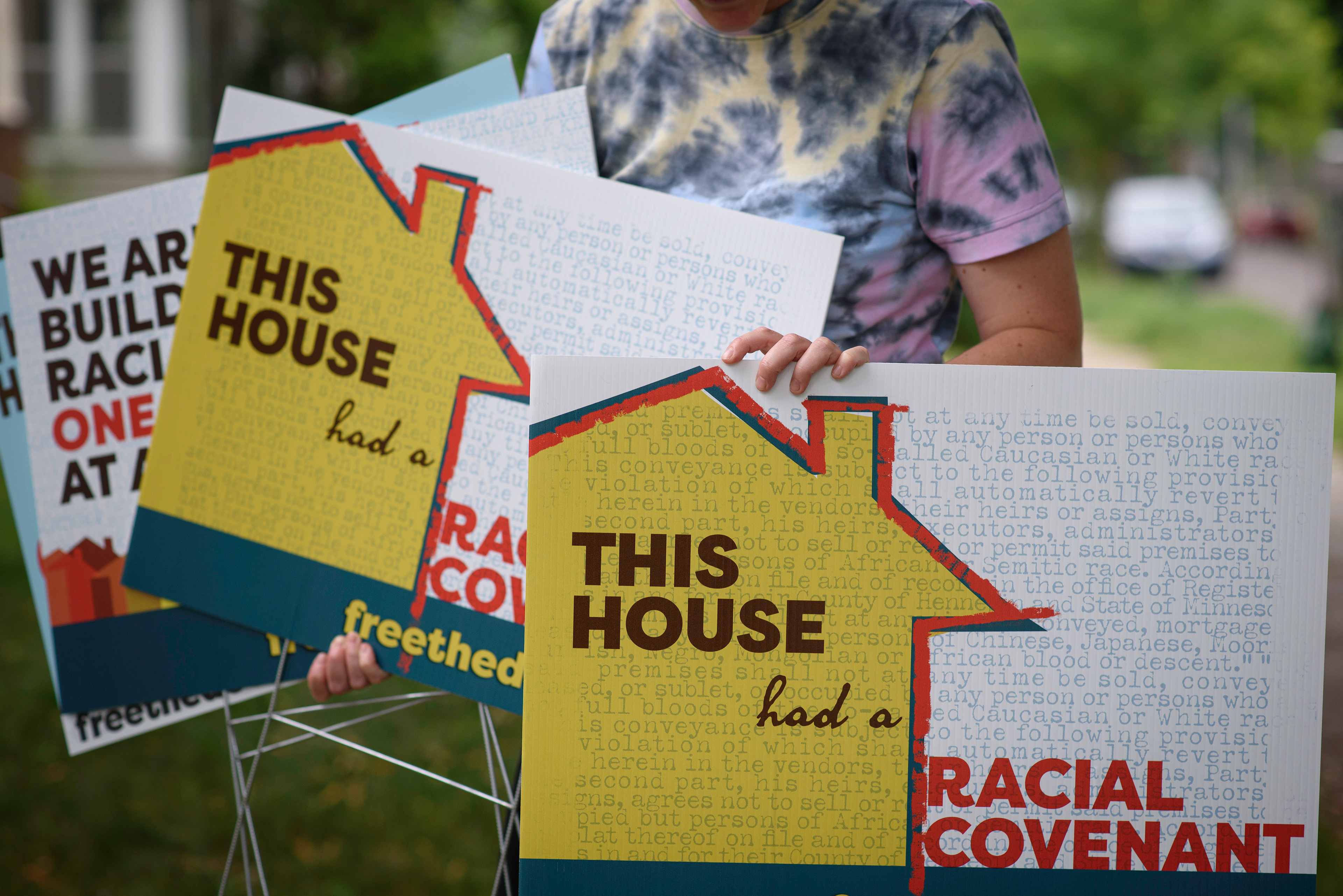 A person in a tye-dyed tshirt holds several yard signs with messages about Free the Deeds.