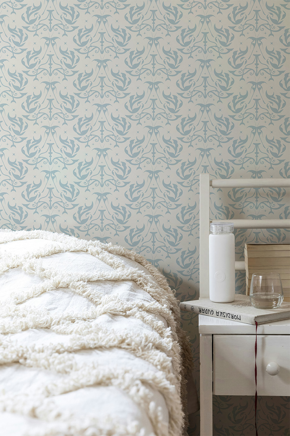 Light blue traditional non-pasted wallpaper with intricate scroll pattern on bedroom walls. White bed with textured cream blanket and white bedside table holding a book, water bottle, and glass.