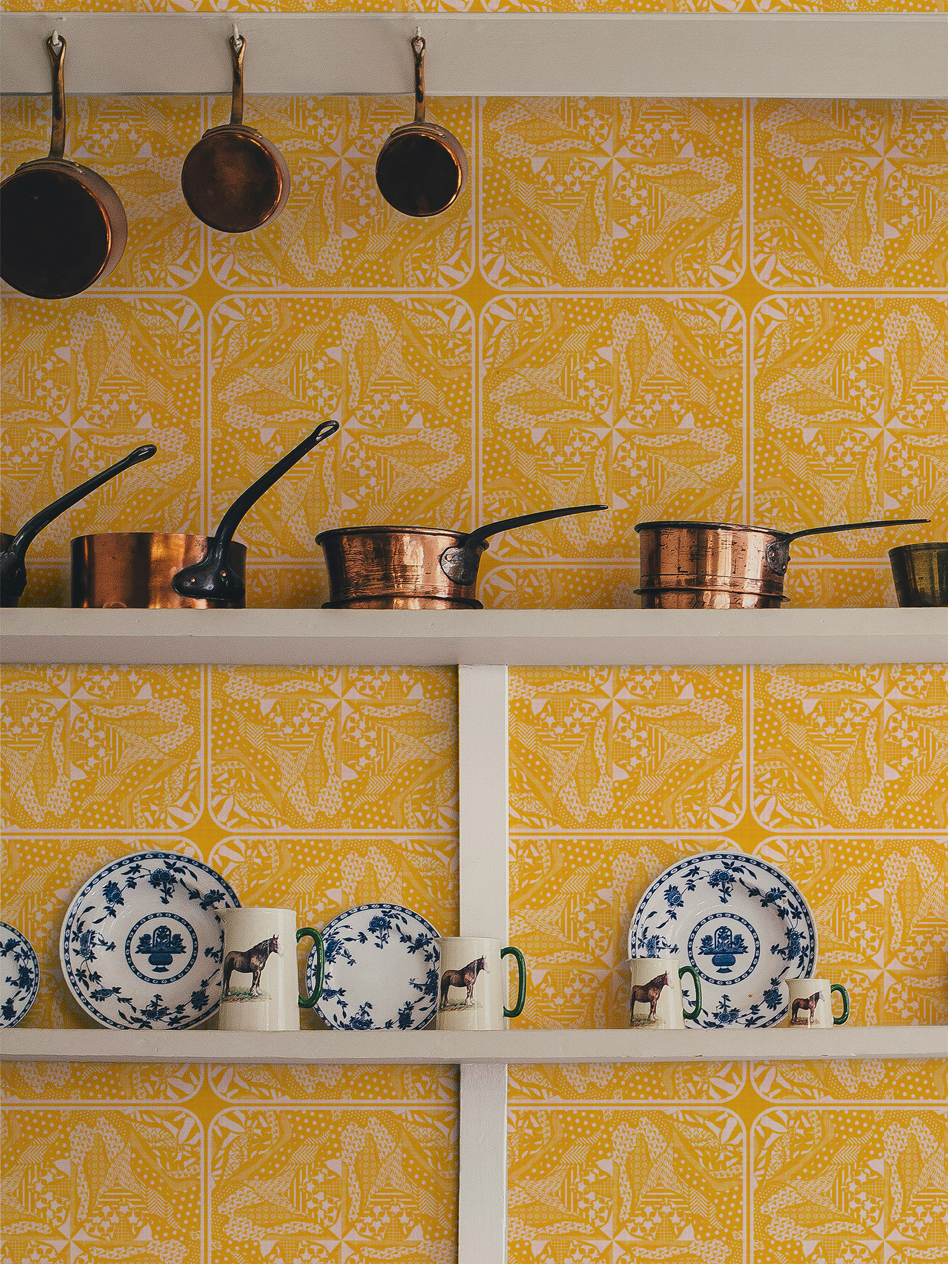 Wallpaper featuring a mustard yellow geometric and organic pattern in a kitchen setting with white open shelving. Shelves display copper pots and blue-and-white floral plates alongside mugs with horse designs, creating a warm vintage kitchen atmosphere.