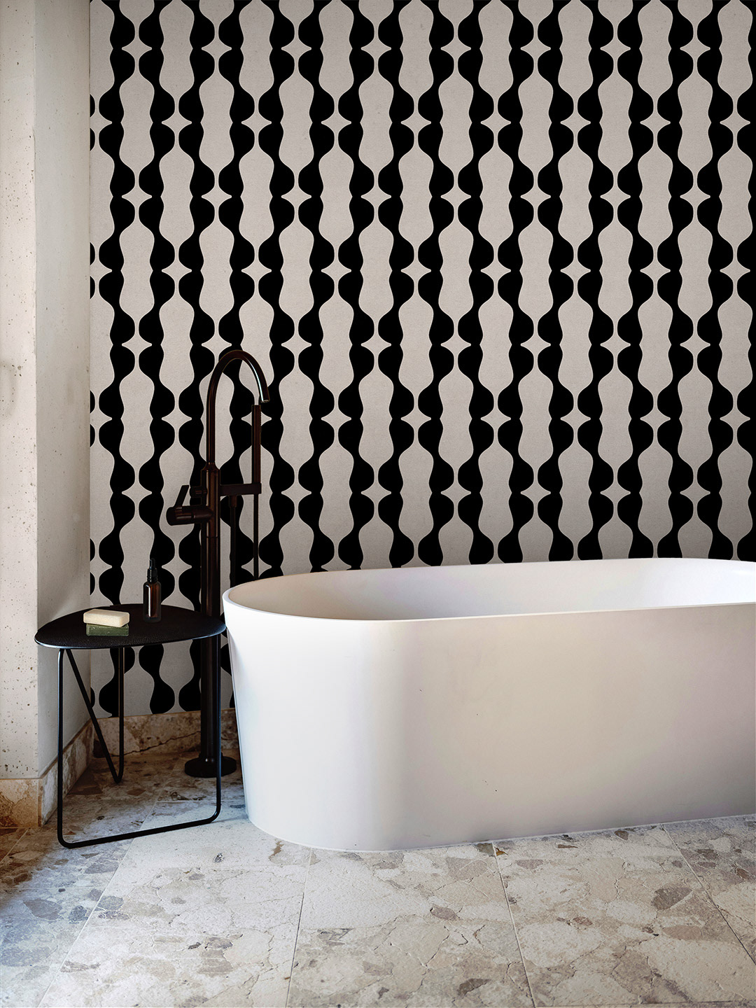 Modern bathroom featuring a black and beige wallpaper with an abstract vertical pattern, paired with a white freestanding bathtub, black faucet, small black side table with soap and bottle, and natural stone tile flooring.