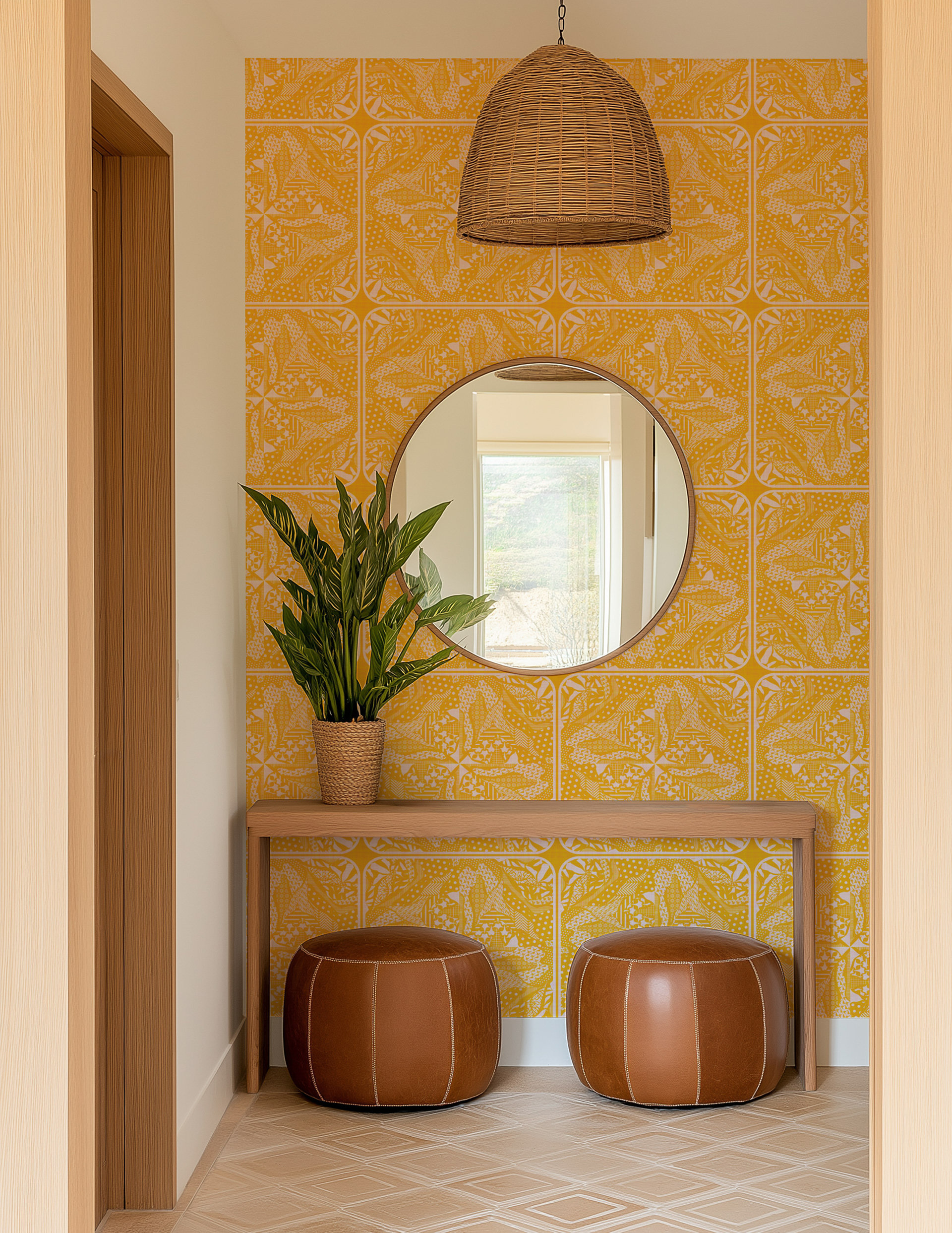 Mustard yellow wallpaper with botanical and organic nature-inspired print on an entryway wall featuring a round mirror, wooden console table, woven plant pot with green leaves, two brown leather poufs, and a hanging woven light fixture.