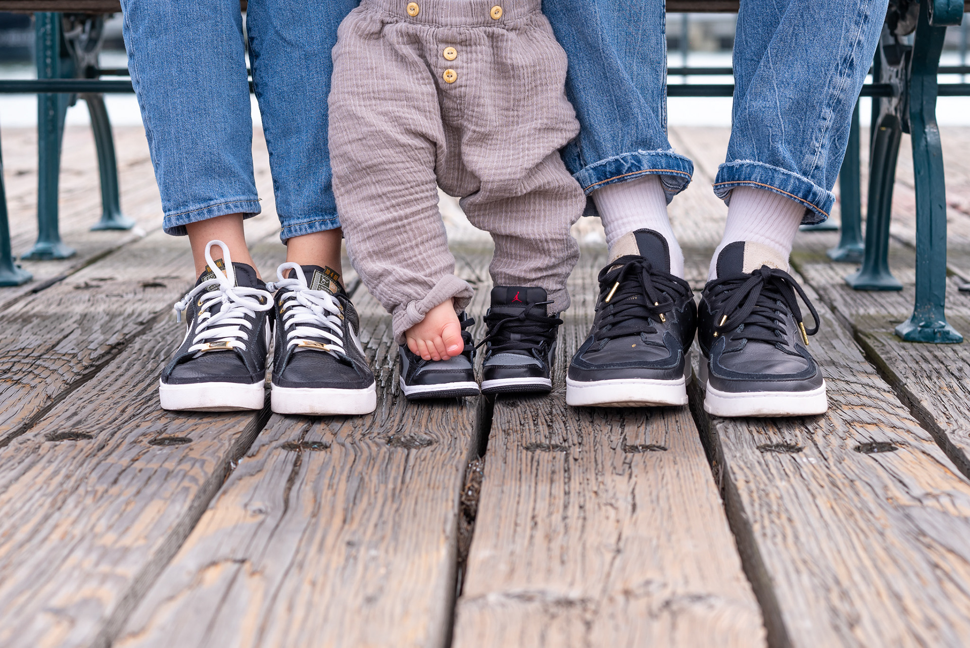 A family session at Pier 7 - mom, dad, and baby boy sitting on a bench with a close-up of their shoes. One of baby's shoes has come off and his foot is peeking out.