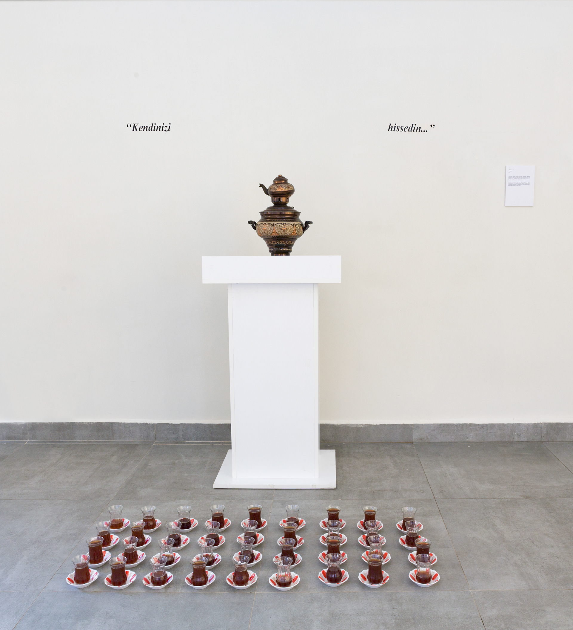  It is Brewing ..., 2017, inlaid copper samovar, white lectern, sticker writing, thin waisted tea cup and traditional red tea plate base (34 pieces). 180x110x120 cm. Independent Art Foundation Art Gallery, Istanbul. Photo: Ismail Coşkun.