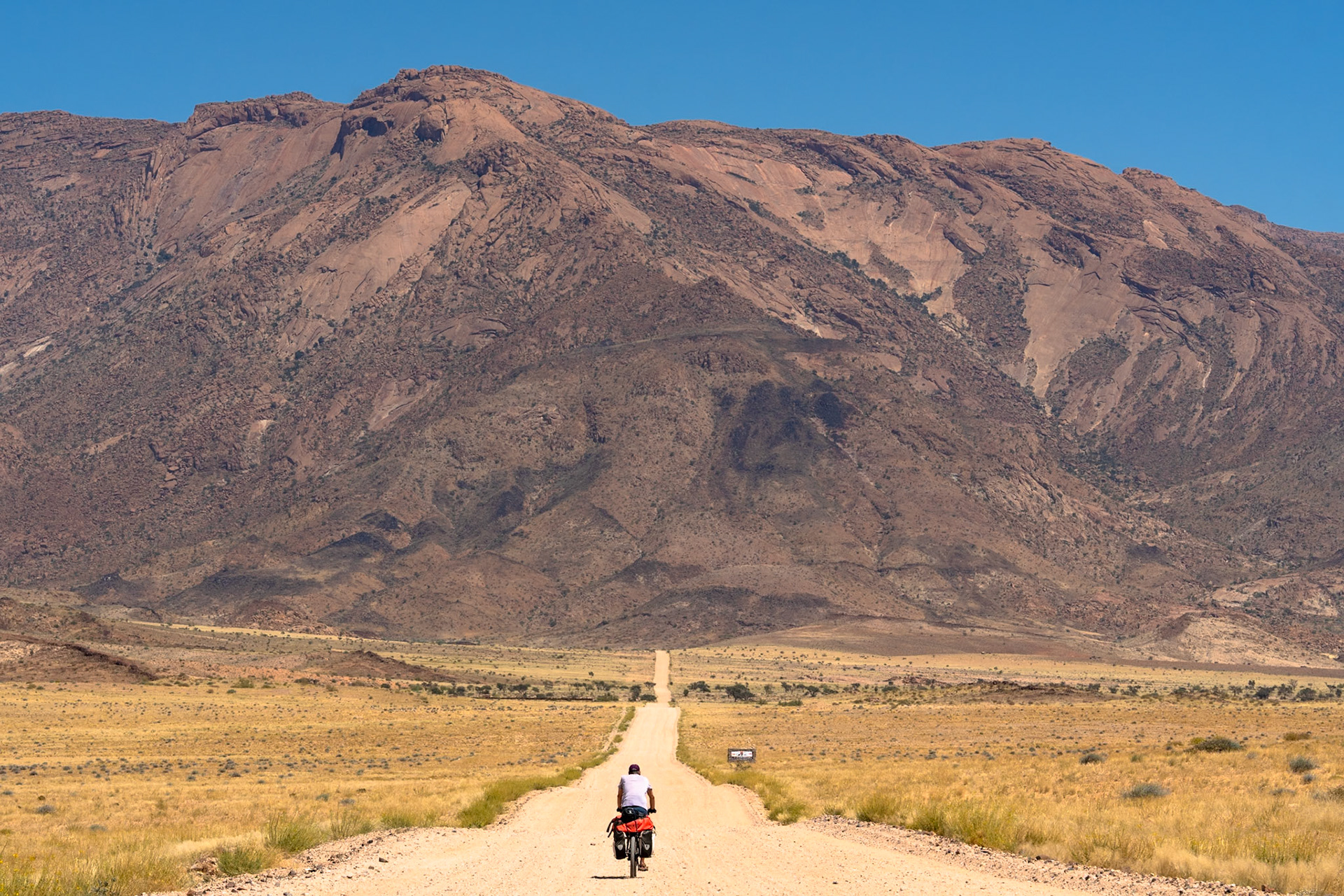 A lone cyclist, seen from behind, rides a touring bicycle with rear panniers and an orange dry bag on a straight, unpaved road. The road traverses a flat, arid landscape with dry, yellowish grass and sparse bushes, leading towards a prominent, massive reddish-brown rocky mountain range under a clear blue sky in the Erongo Region of Namibia. The mountain exhibits visible geological formations and scattered dark vegetation. Spitzkoppe, Namibia.