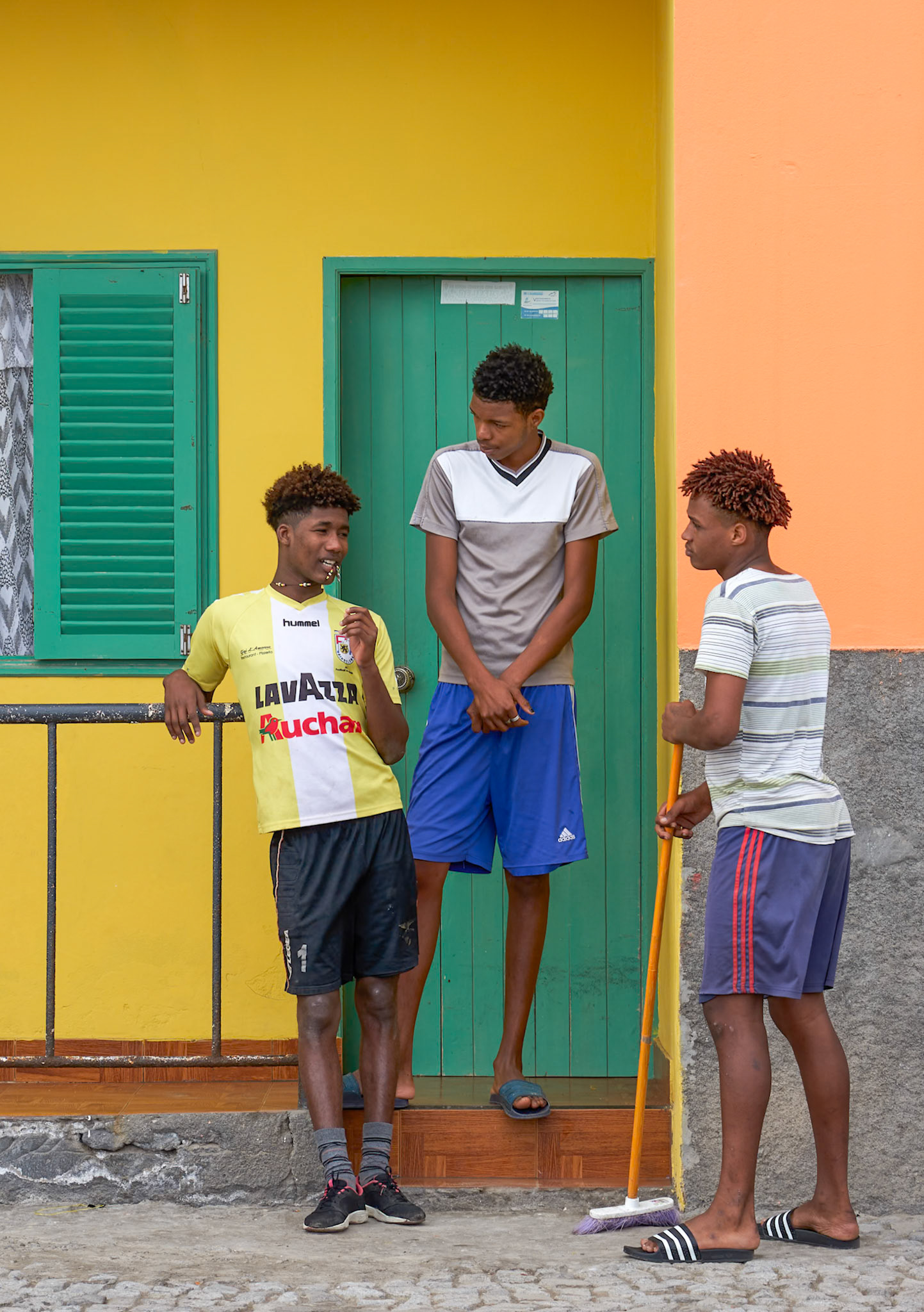 Three young men stand in front of a brightly colored building in a tropical location. The building has a yellow facade with green shutters on a window and a green door. The man on the left leans on a railing, wearing a yellow and white sports jersey and black shorts. The man in the center wears a grey and white t-shirt and blue shorts. The man on the right holds a broom and wears a striped t-shirt and blue shorts. The ground is paved with stone and concrete. The lighting suggests it is daytime.