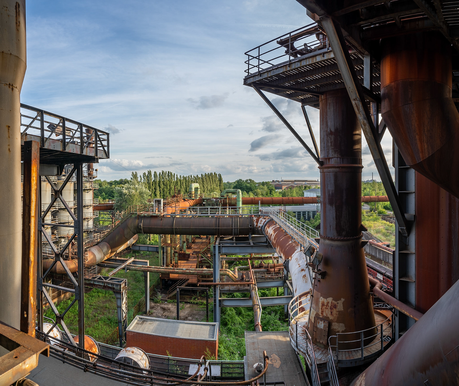 An expansive view of an abandoned industrial complex, featuring large, rusted metal pipes, walkways, and structural elements under a partly cloudy sky. The scene is set in the Ruhr area of Germany, with overgrown vegetation encroaching on the industrial structures. The lighting suggests late afternoon or early evening.