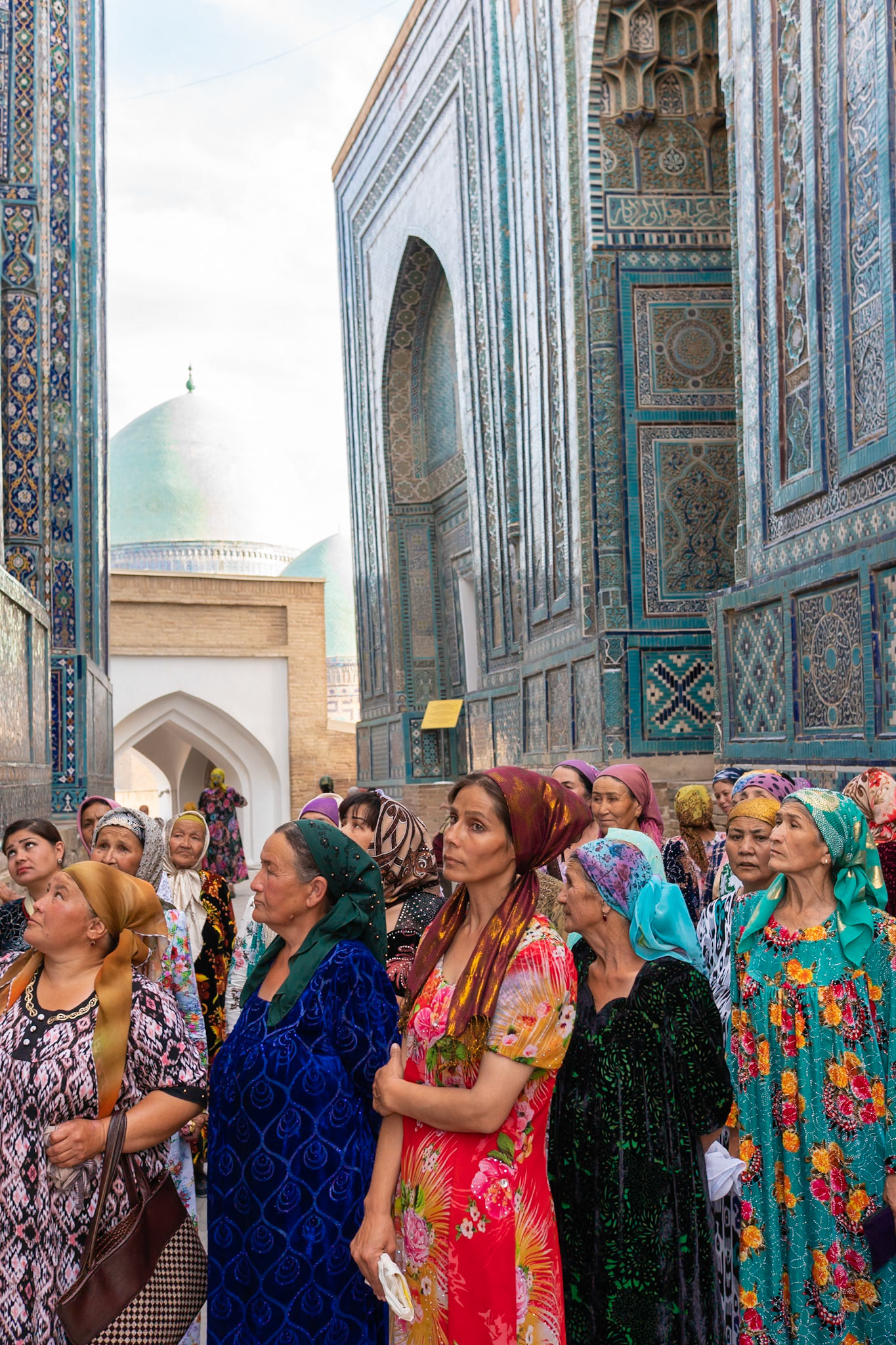 A group of women, adorned in vibrant, patterned headscarves and dresses, stand amidst the stunning turquoise-tiled architecture of a historical site in Samarkand, Uzbekistan. The composition focuses on their faces, turned upwards and outwards with expressions of contemplation and awe, set against the intricate geometric and calligraphic details of the mausoleums. Soft daylight illuminates the scene, highlighting the rich textures and colors of both the women's attire and the ancient structures, creating a mood of reverence and cultural immersion.