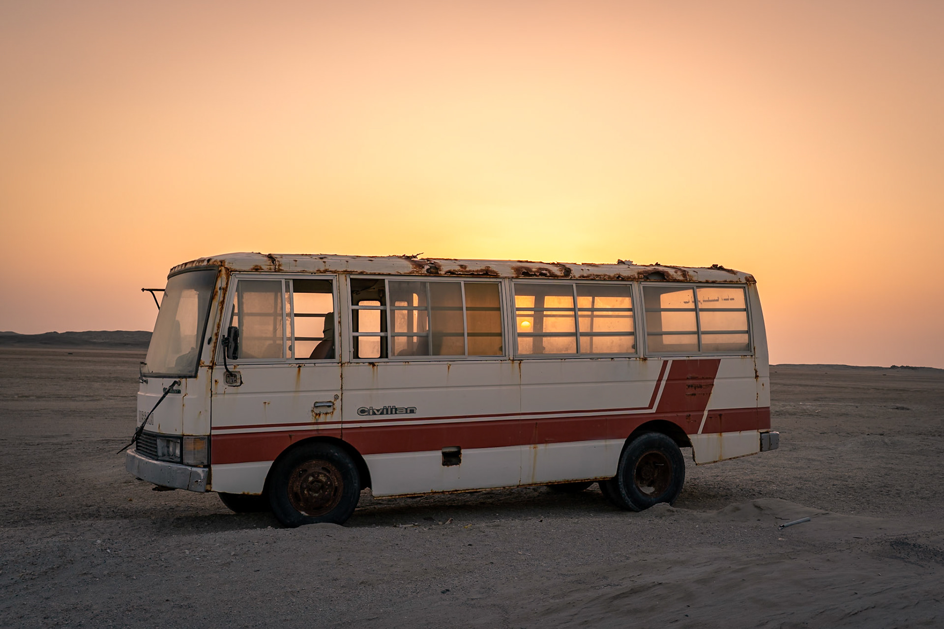 A rusty, abandoned white and red civilian bus sits in a barren, sandy landscape under a warm, orange sunset sky. The sun is visible through the bus windows, reflecting off the dusty glass. The ground is uneven with sand dunes and sparse gravel. The location is in the desert region of Oman.