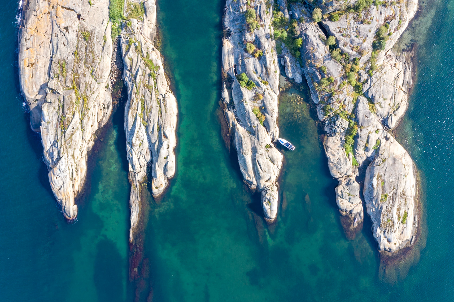 An aerial view shows several rocky islets with sparse vegetation, jutting out of clear, turquoise water. A white boat with solar panels is moored in a narrow channel between two of the islets. The sun casts shadows on the textured surfaces of the rocks, highlighting their contours and crevices. The scene is located in the coastal region of Southern Norway.
