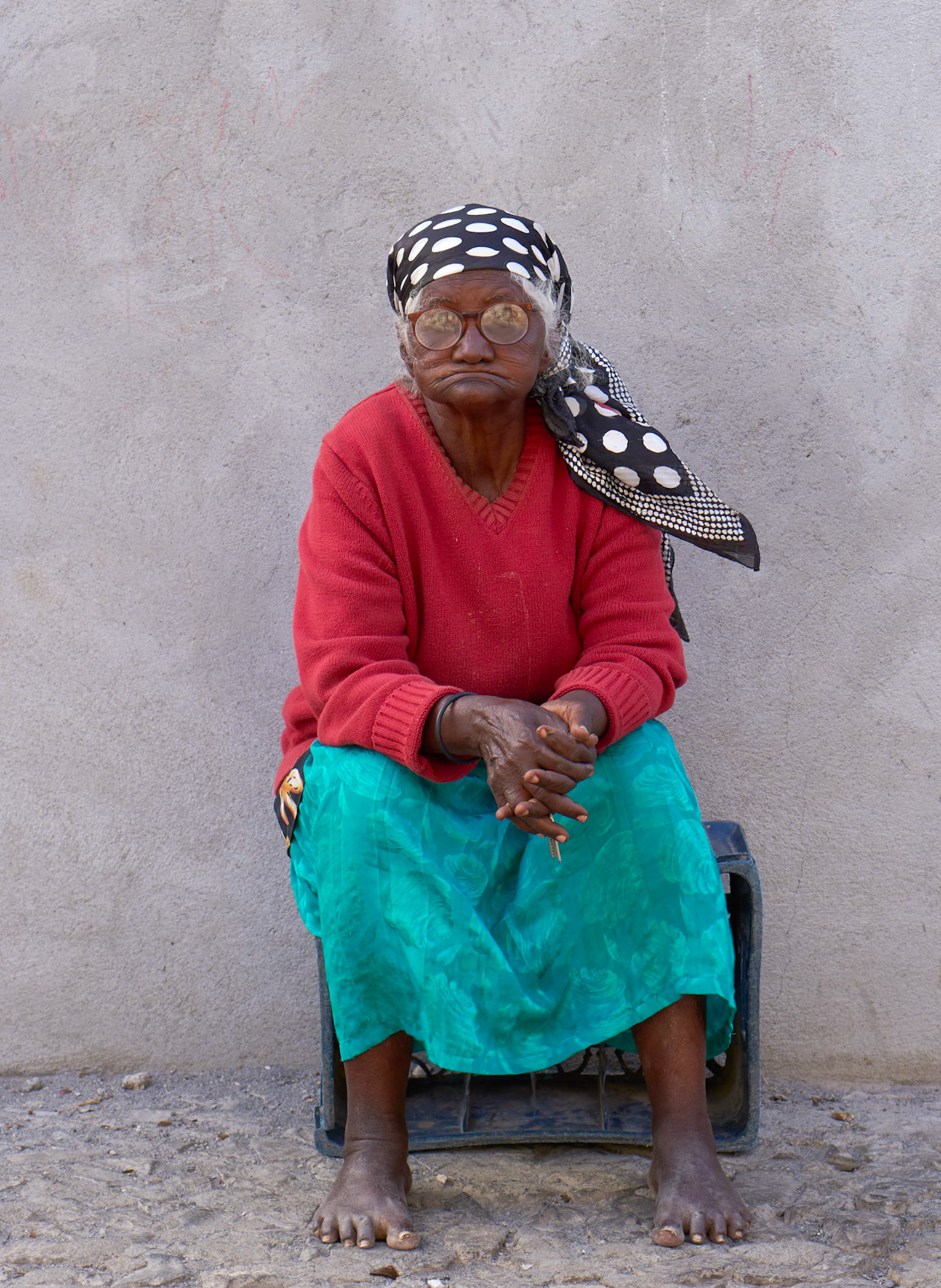 An elderly woman with glasses and a black and white polka dot headscarf sits in front of a textured concrete wall in what appears to be a rural area of southwestern Mauritania. She wears a red V-neck sweater and a bright turquoise skirt. Her hands are clasped in her lap, and she is barefoot. The ground is rough and stony.
