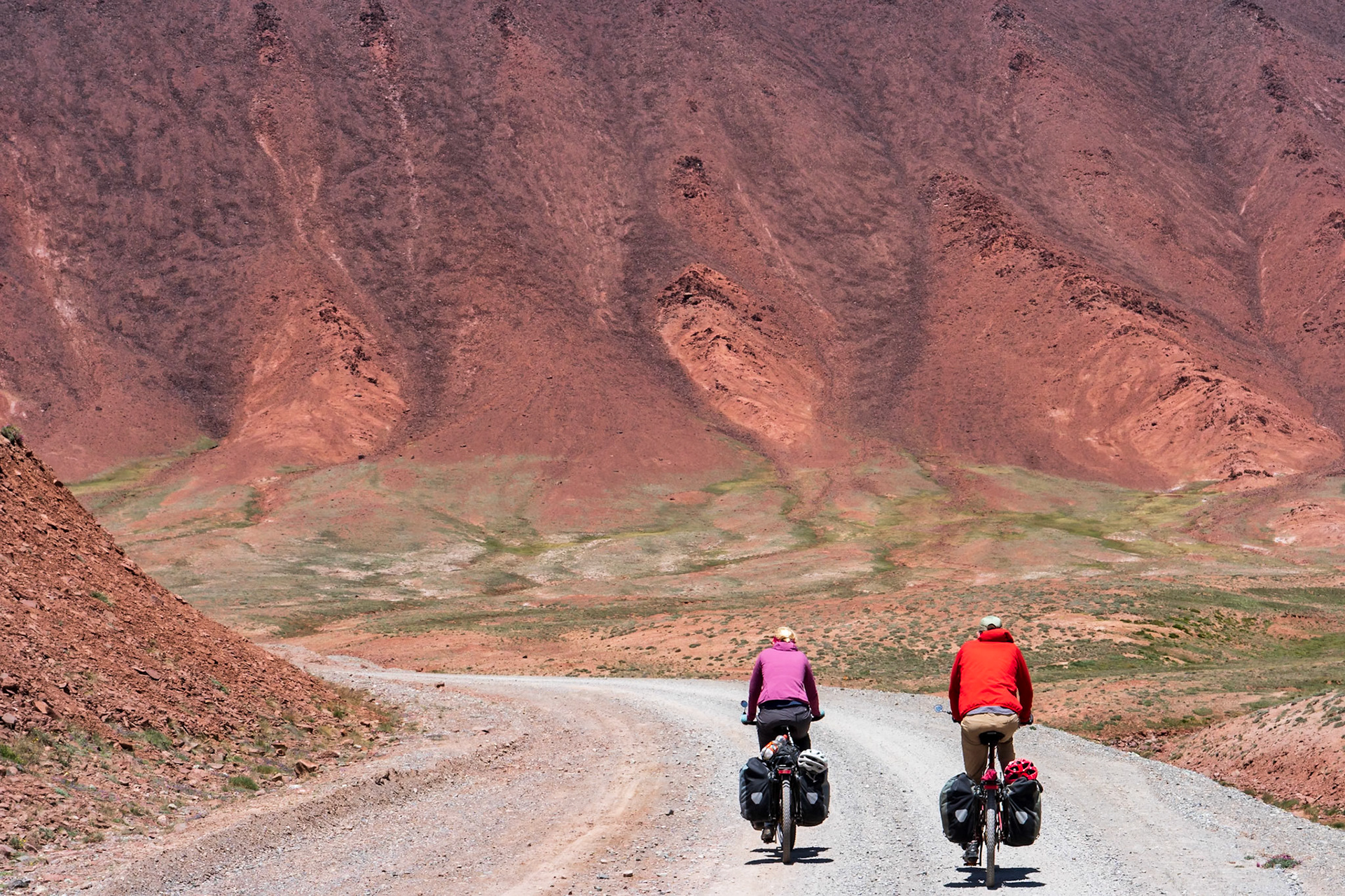 Two cyclists, laden with panniers, pedal along a dusty, winding road through a vast, arid landscape. The stark, red-hued mountains rise dramatically in the background, their textured slopes carved by time and erosion. Patches of sparse green vegetation dot the valley floor, contrasting with the dominant earthy tones. The bright sunlight casts sharp shadows, emphasizing the rugged terrain and creating a mood of adventurous solitude. The composition draws the eye along the road, inviting the viewer into the expansive, remote scenery, likely in a high-altitude desert region of Central Asia.