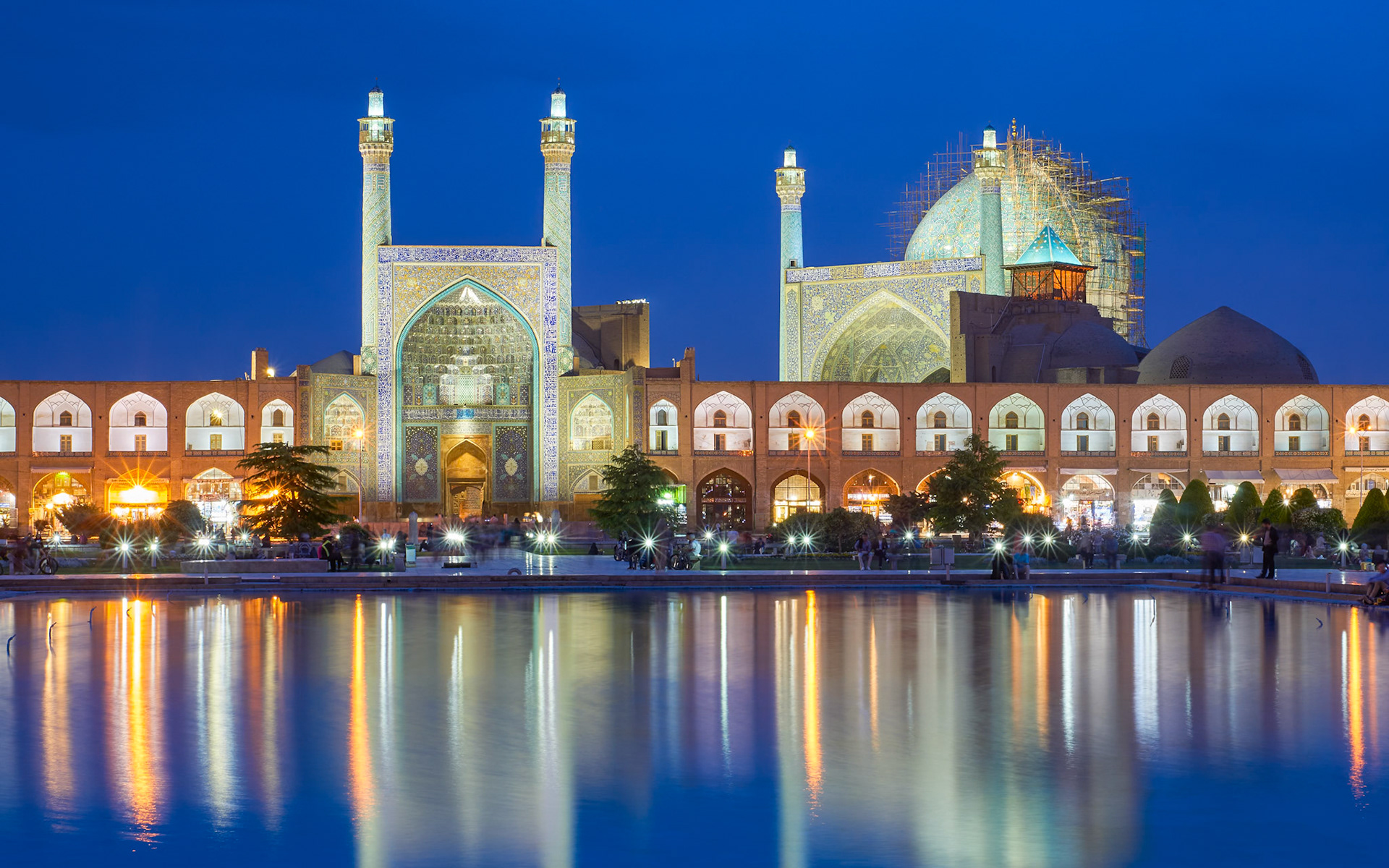 A wide-angle, long-exposure photograph captures the Imam Mosque in Isfahan, Iran, at dusk. The mosque's facade, adorned with intricate blue and yellow tilework, is illuminated by artificial lights. Two tall minarets flank the central dome, which is partially covered in scaffolding, indicating ongoing restoration. In front of the mosque, a long, arched arcade with shops is visible, also lit from within. A large rectangular pool in the foreground reflects the lights of the mosque and arcade, creating a shimmering effect on the water's surface. The sky is a deep twilight blue. Several people are seated or walking along the edge of the pool and in front of the arcade.