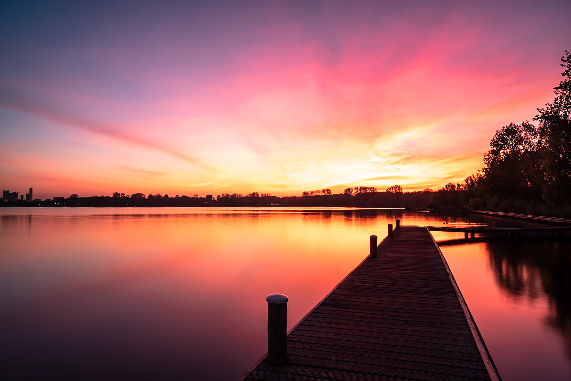 A wooden pier extends into a tranquil lake reflecting the vibrant hues of a dramatic sunset. The sky transitions from deep purple and pink overhead to fiery orange and yellow near the horizon, silhouetting a distant city skyline and trees along the far shore. The calm water mirrors the sky's spectacle, creating a serene and breathtaking atmosphere. This scene captures the beauty of twilight near Rotterdam, Netherlands.