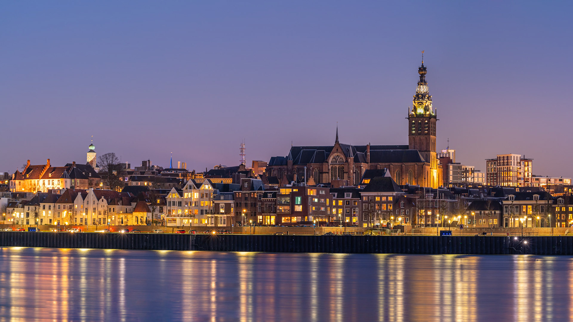A panoramic view of the Nijmegen skyline unfolds under a twilight sky, with the imposing Grote Kerk dominating the center. Warm lights spill from the windows of historic buildings and modern apartments, reflecting on the calm water in the foreground. The composition captures the transition from day to night, creating a serene and inviting atmosphere in this Dutch city.