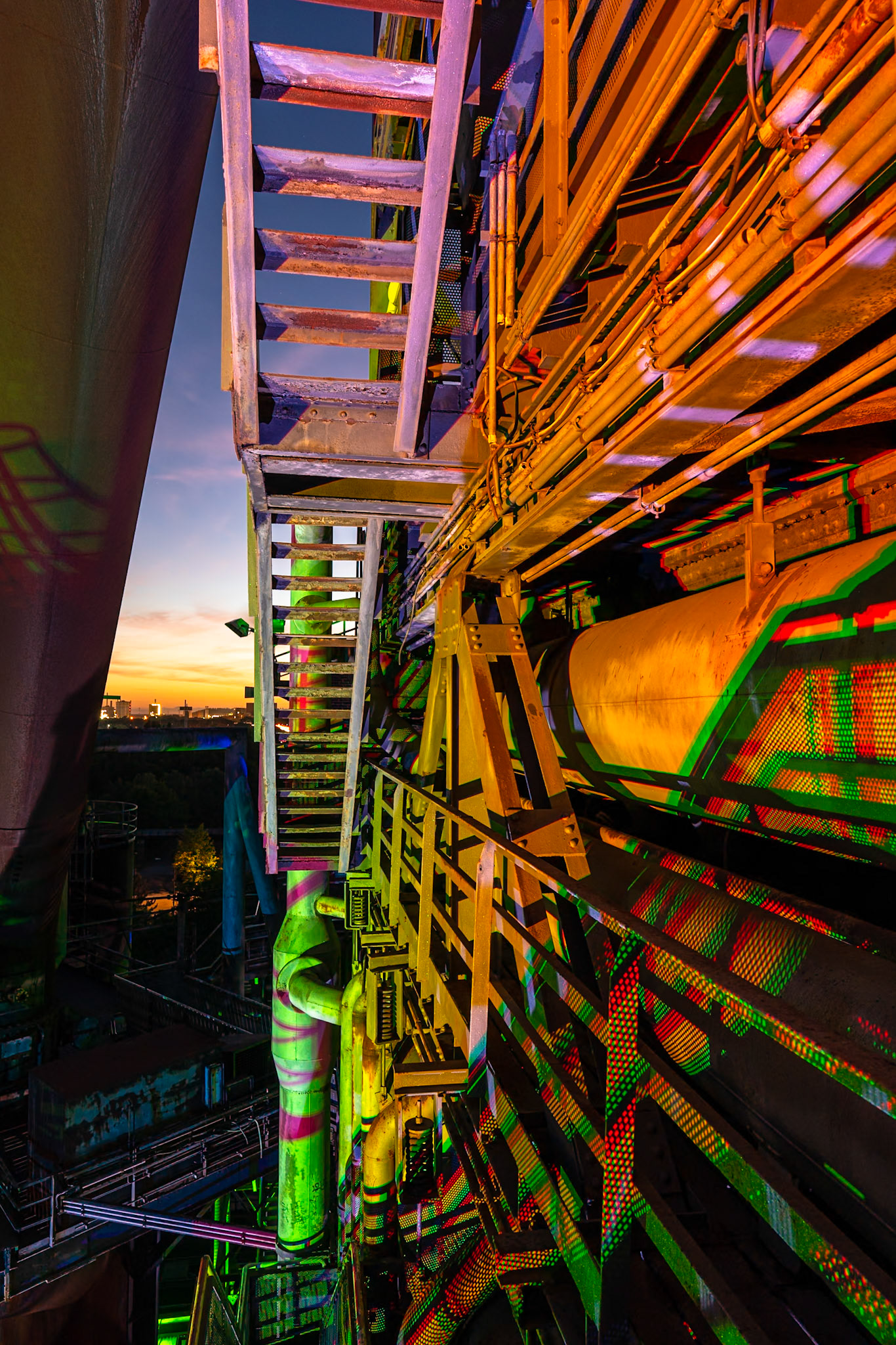 A complex industrial structure with metal stairs, pipes, and beams is illuminated by colorful lights, casting patterned shadows. The scene is set against a twilight sky with a distant cityscape visible. The location is in the Ruhr Area, Germany.