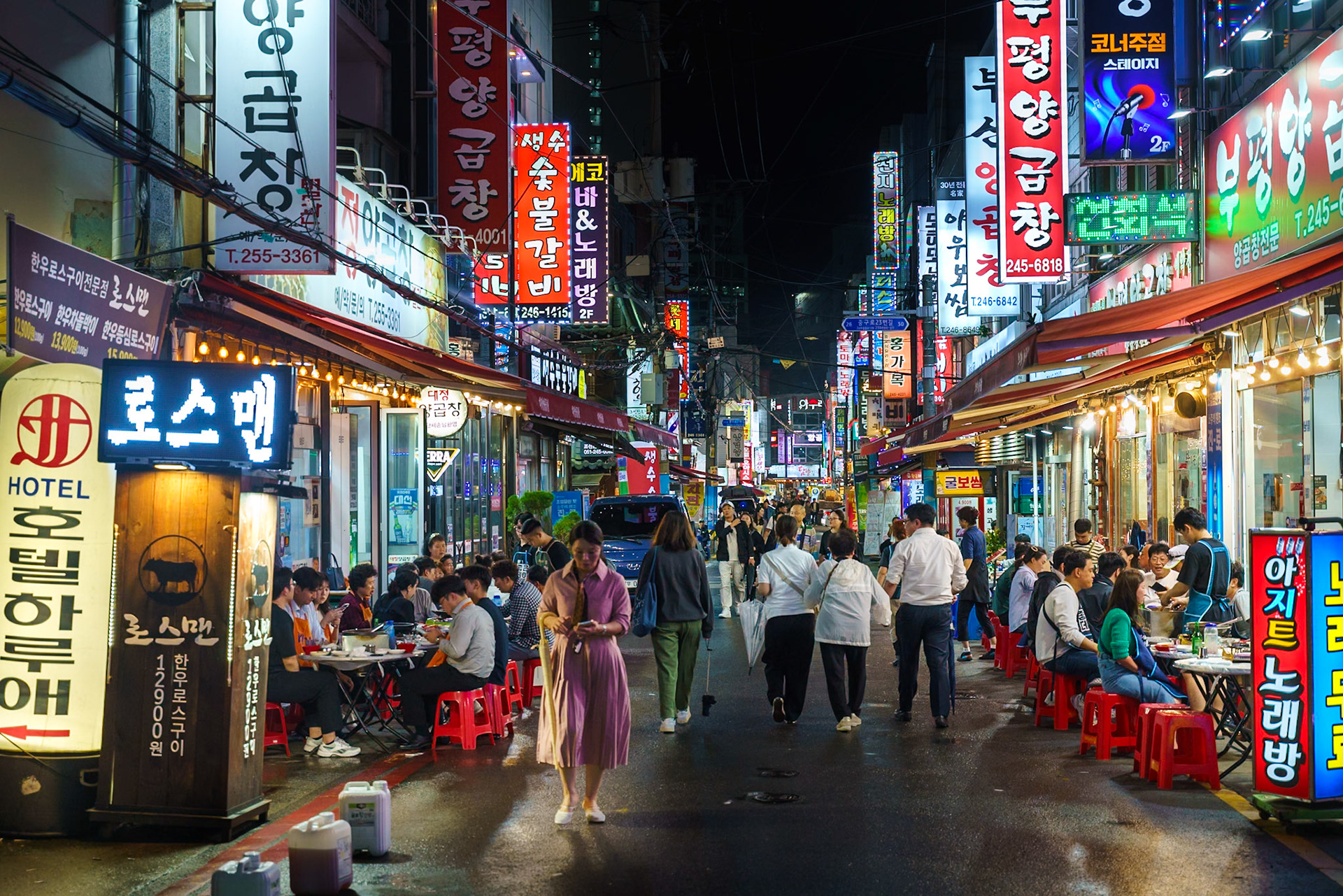 A bustling street scene in South Korea at night, illuminated by a vibrant array of neon signs and string lights. People are gathered at outdoor eateries, seated on red stools around tables laden with food, creating a lively and convivial atmosphere. The composition draws the eye down the narrow street, flanked by shops and restaurants, with a dense network of electrical wires overhead adding to the urban texture. The mood is energetic and inviting, capturing the essence of a lively night market.