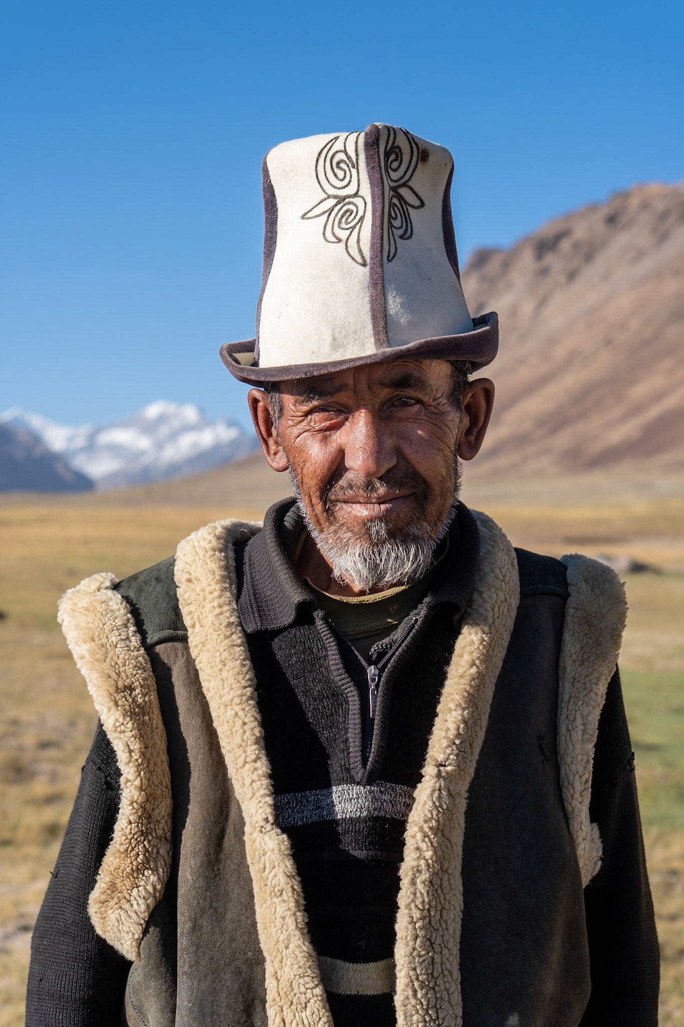 A medium shot portrait of an elderly man wearing a traditional white and brown embroidered hat and a shearling-lined vest over a dark sweater. He has weathered skin and a grey beard. The background shows a dry, grassy plain with distant, snow-capped mountains under a clear blue sky. The lighting suggests it is daytime with the sun casting shadows on the man's face.