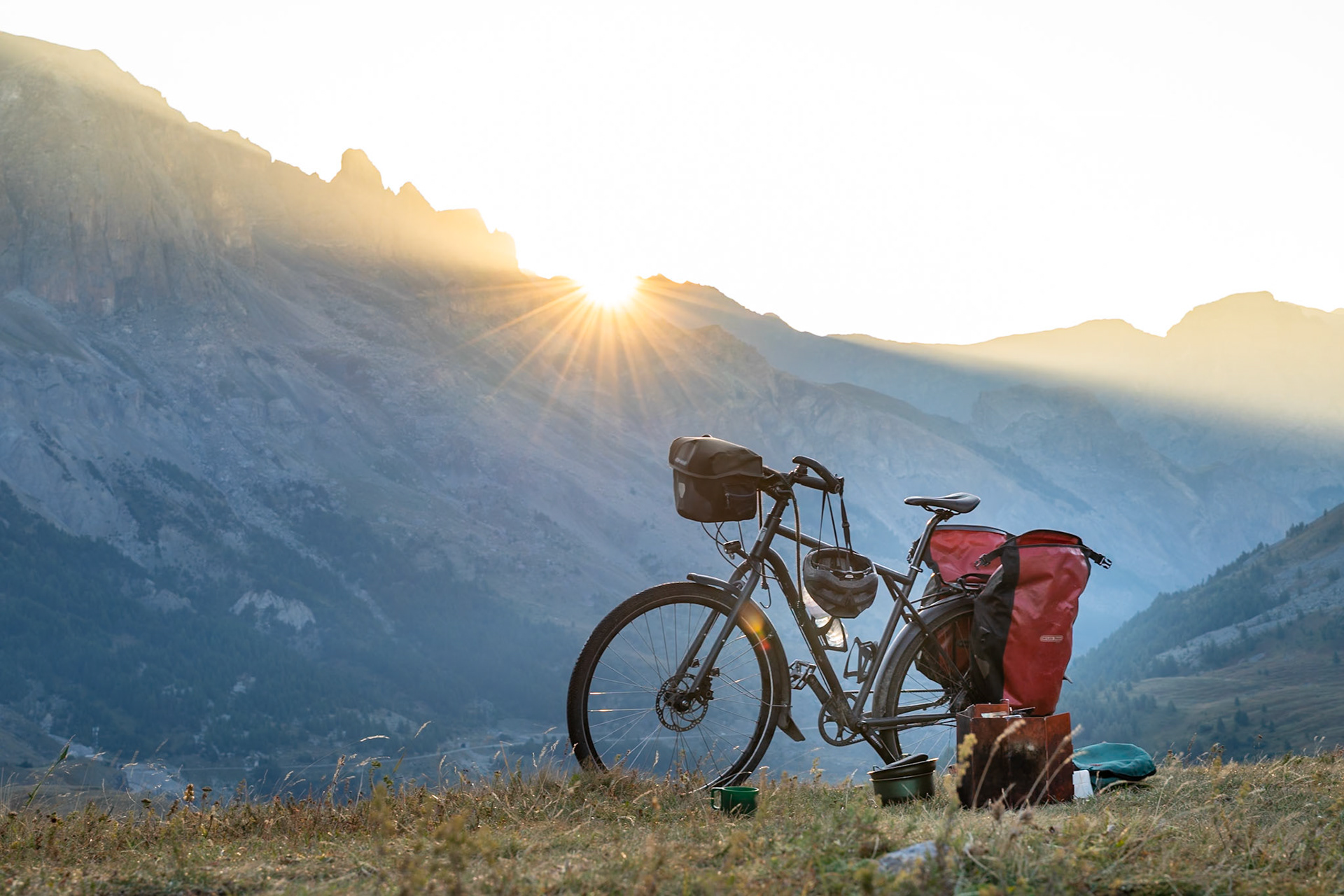 A touring bicycle equipped with panniers and a helmet is parked on a grassy slope in the mountains, with the sun setting behind jagged peaks. The low sun casts long rays across the rugged landscape. The scene is located in the French Alps.